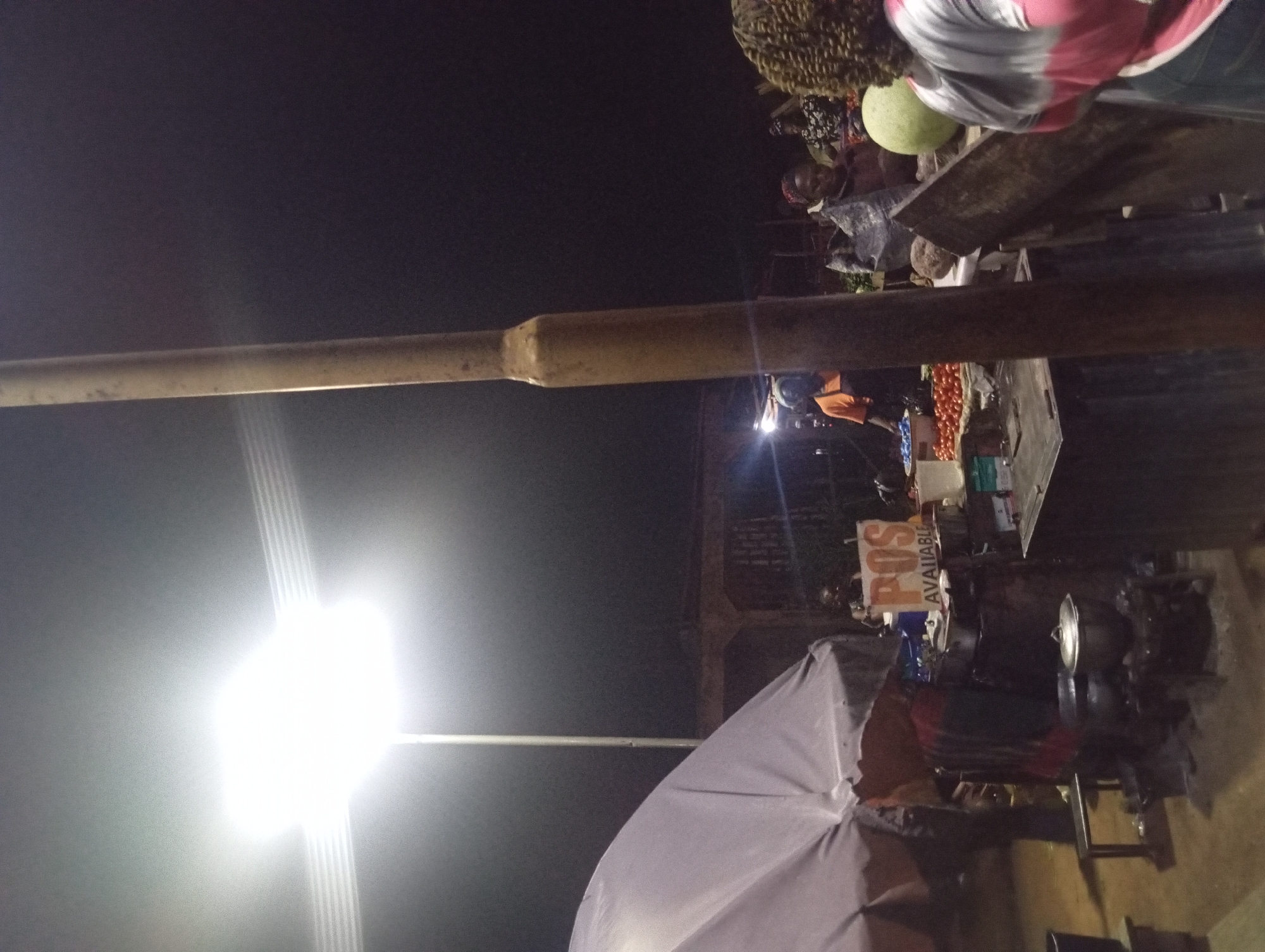 An outdoor night market scene unfolds under a bright street lamp in Unknown, Unknown. The foreground is dominated by a weathered metal pole. To the left, a large, pale umbrella partially obscures a stall where cooking pots are visible. A sign with "POS AVAILABLE" written in white on an orange background is displayed on the stall. In the background, several people are gathered around other stalls. One stall features a display of tomatoes, and a person in an orange shirt is visible behind it. Further to the right, more individuals are present, with a large green melon and some produce on display. The overall atmosphere suggests a busy marketplace operating after dark.