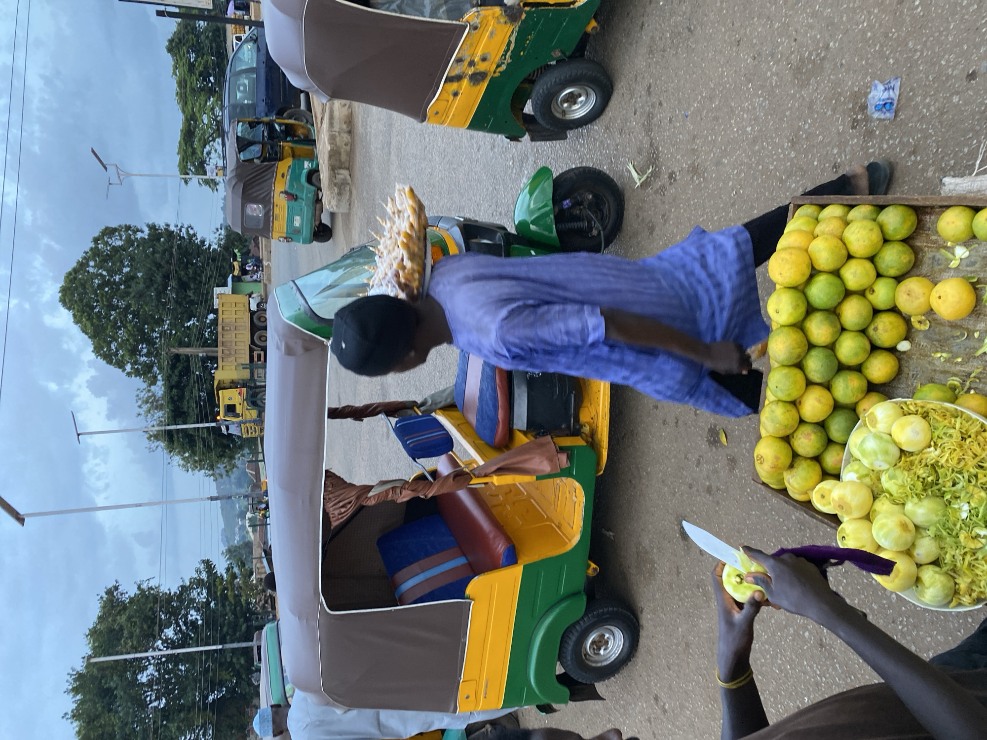 The image depicts a street scene in Bukuru, Nigeria, under an overcast sky. In the foreground, a person is preparing a pile of what appears to be green fruit, possibly guavas, with a knife. Adjacent to this, a wooden crate is filled with a large quantity of oranges, some green and some yellow. To the left, partially visible, is another person wearing a brown shirt and a yellow bracelet on their left wrist.
Dominating the midground are several three-wheeled auto-rickshaws, known locally as "keke napep." One prominent keke napep, painted green and yellow with a brown canopy, has its doors open, revealing blue and red striped seating. A person in a blue, patterned tunic is walking away from this keke, carrying a tray of what looks like fried snacks on their shoulder. Another keke, also green and yellow, is parked to the right, its canopy partially obscuring the vehicle. Further in the background, a yellow dump truck is visible, along with other vehicles, including a dark-colored minivan. The environment appears to be an open area, possibly a market or a roadside, with sparse vegetation and buildings in the distance. Streetlights are also present, indicating a developed urban area. The ground is paved with asphalt.