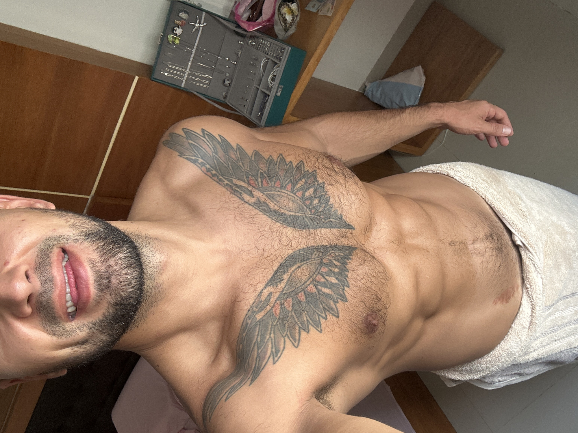 A shirtless man with a beard and a winged tattoo on his chest stands in a room, wearing a towel around his waist. He is smiling with his mouth slightly open. To his right, on a wooden surface, is an open jewelry box containing various necklaces and rings, as well as a small decorative item. In the background, a wooden wall and part of a bed are visible. The image appears to be a personal photograph taken by the subject himself. There is no indication that this image is located in Nha Trang Ward, Vietnam.