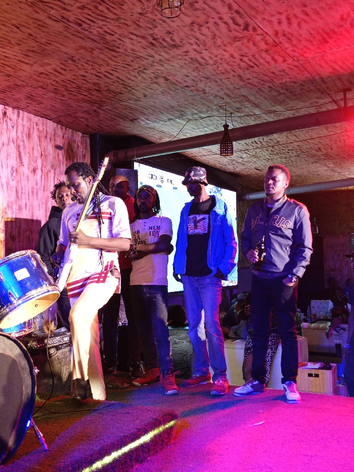 The image captures an indoor performance venue in Nairobi, Kenya. Six adult males are positioned on or immediately adjacent to a stage, illuminated by prominent red and purple stage lighting.
In the foreground, a man with dark dreadlocks, dressed in a white tunic with distinct red horizontal stripes and light-colored trousers, holds and is oriented to play a light-colored electric guitar. A blue tom-tom drum, part of a larger drum kit, is visible on the stage to his immediate left.
Behind the guitarist, positioned from left to right, are four additional men. The first, partially obscured, is dressed in dark attire. The second, also partially visible, wears a red shirt. The third, with dark dreadlocks, wears a white t-shirt and has his arms crossed. The fourth wears a camouflage bucket hat, a blue jacket, and blue jeans, over a dark graphic t-shirt. On the far right of the frame, a sixth man stands, wearing a grey bomber jacket with a prominent "PARIS" or similar logo, dark trousers, and holds a brown glass bottle in his right hand.
The stage floor is dark and appears to be carpeted. The background features light-toned wooden wall paneling on the left, a textured ceiling, and two visible hanging light fixtures with woven shades. A large, illuminated screen displaying white graphical elements on a dark background is positioned centrally behind the group. The environment indicates a dedicated space for live entertainment or a social gathering within the city.