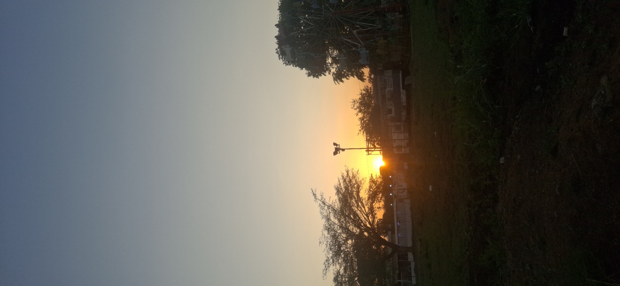 The image captures a sunset scene in Tinaquillo, Venezuela. The sky transitions from blue to orange as the sun sets on the horizon. In the foreground, there is a grassy area with trees and a building in the background. A lamppost stands tall, silhouetted against the bright sunlight. A Ferris wheel can be seen in the distance to the right.
