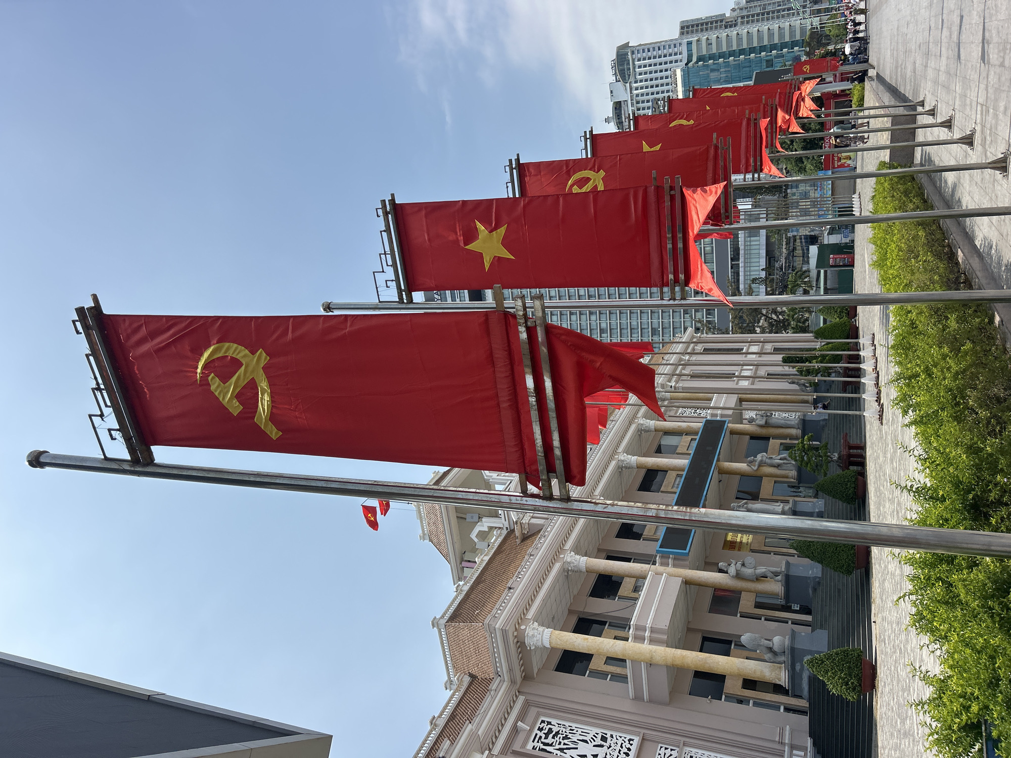 This image, rotated 90 degrees counter-clockwise from its original orientation, depicts a patriotic display along an urban street. A prominent row of red flags, mounted on silver metallic poles, lines what appears to be a wide pedestrian walkway. These flags feature two main designs: some display the gold hammer and sickle symbol of the Communist Party, while others show a single gold five-pointed star, representing the national flag of Vietnam.
In the mid-ground, behind the flags, stands a stately, classical-style building with multiple stories, adorned with large pillars, statues, and decorative architecture. Beyond this building, a modern city skyline emerges, characterized by numerous high-rise buildings with glass facades. The ground level includes a paved area and well-maintained green bushes.
The sky is clear and bright blue, suggesting a sunny day. In the far distance along the walkway, a few indistinct figures can be seen, indicating public activity. The presence of both the Vietnamese national flag and the Communist Party flag strongly indicates a location in Vietnam, specifically Nha Trang, as provided. On a sign partially obscured in the background, the word "MIX" is faintly visible. Other text on signs on the classical building is present but illegible. The overall scene conveys a sense of national pride and urban development.