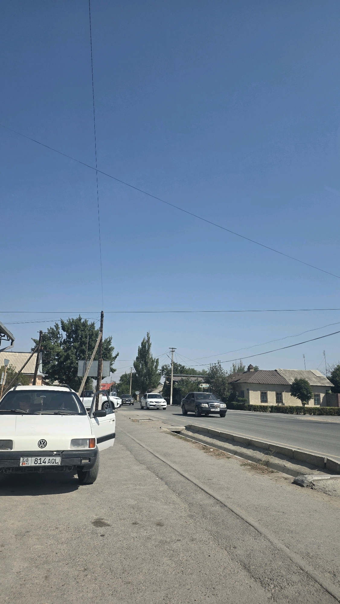 The image shows a street scene in Tokmok City, Kyrgyzstan, on a clear, sunny day. A white Volkswagen car is parked on the left side of the road, with its driver's side door open. The car has a license plate that reads "08 814 AQL". In the background, several other vehicles are driving on the asphalt road. These include a white sedan, a dark-colored sedan, and another white car further down the road. On the right side of the road, there are houses with beige walls and brown roofs. Trees are visible along the street and behind the houses. Power lines are strung across the sky, supported by wooden poles. The street appears to be a main thoroughfare in the city.