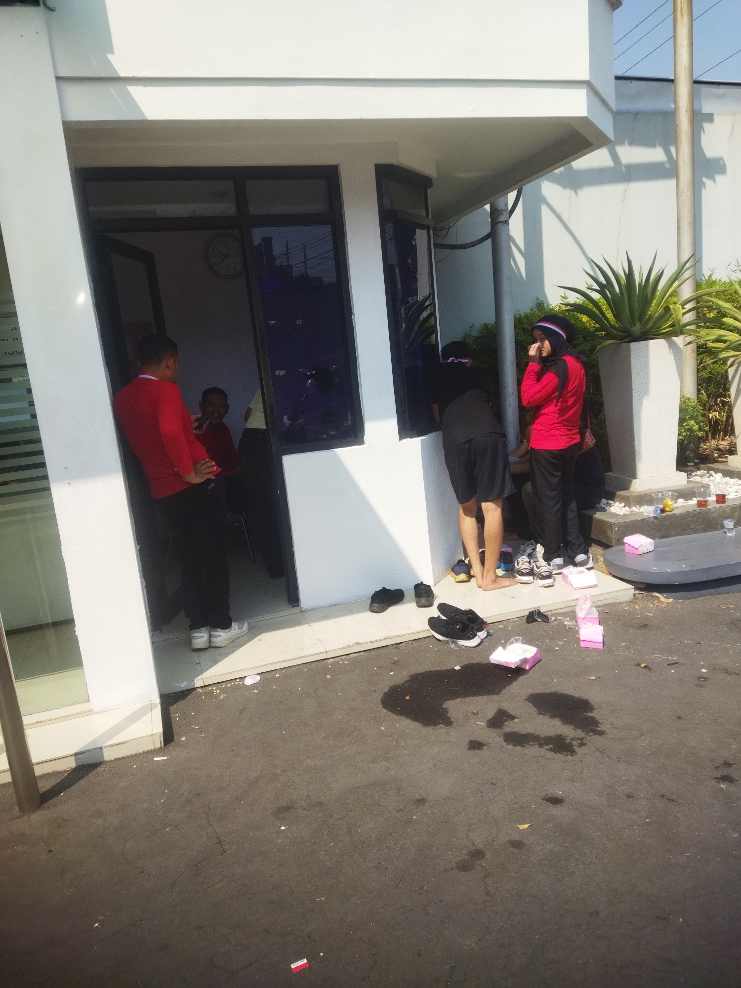 A group of people, some wearing athletic attire and headscarves, are gathered outside a building with white walls and large glass windows. One individual in a red long-sleeved shirt and black pants stands by an open doorway, facing inwards and appearing to speak with someone inside. Another person in black shorts and a black t-shirt is partially visible from behind, facing away from the camera, and is barefoot. A woman wearing a red and black outfit with a black headscarf is standing to the right, looking down and touching her face. Several pairs of shoes, including sneakers and black slip-on shoes, are scattered on the ground near the building's entrance, as well as discarded pink boxes and cups. The ground is paved with asphalt, and there are puddles of liquid in some areas. A tall pole stands to the right of the building, and a potted plant with spiky green leaves is visible behind the people. The scene appears to be outdoors in a sunny environment. The presence of a clock inside the building and the architectural style suggest a location within Wonogiri, Indonesia, potentially an office or facility.