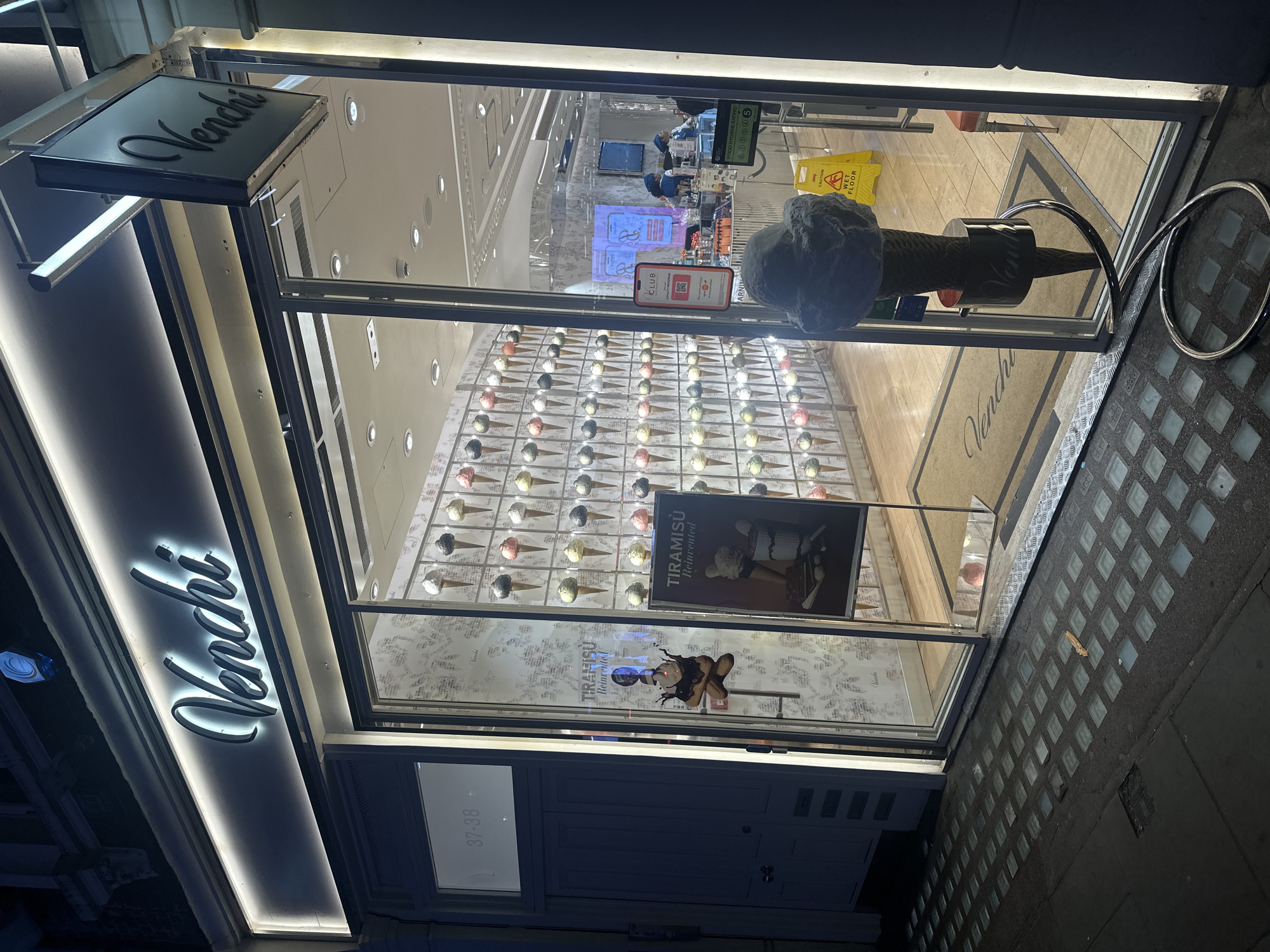 The image captures the illuminated storefront of a Venchi shop at night, likely in London, United Kingdom, given the provided context and visible cues. The scene is dark outside, emphasizing the brightly lit interior and exterior signage.
The shop's name, "Venchi," is prominently displayed in two places: a rectangular hanging sign and a larger, backlit sign on the building's facade, both using a distinctive script font. The address numbers "37-38" are visible on the lower left of the facade.
Through the large glass windows, the shop's interior is visible. A striking feature is a wall adorned with numerous decorative ice cream cones of various colors and flavors, arranged in a grid pattern. Near the entrance, a large, dark-colored, sculptural ice cream cone stands as a prominent display. Posters advertising "TIRAMISÙ Reinvented" are also visible inside the window.
Further inside, what appears to be a counter area can be seen, along with a yellow "WET FLOOR" caution sign on the floor. A "FOOD HYGIENE RATING 5" sticker is affixed to the window, indicating excellent food safety standards. A digital display or poster shows a "CLUB" loyalty program with a QR code.
The pavement outside the shop features distinctive square glass blocks embedded into the dark surface, a common sight in London. A metallic, ring-shaped street furniture object is partially visible on the right. The overall impression is of a clean, well-maintained, and inviting gelato and chocolate shop open for business in the evening.