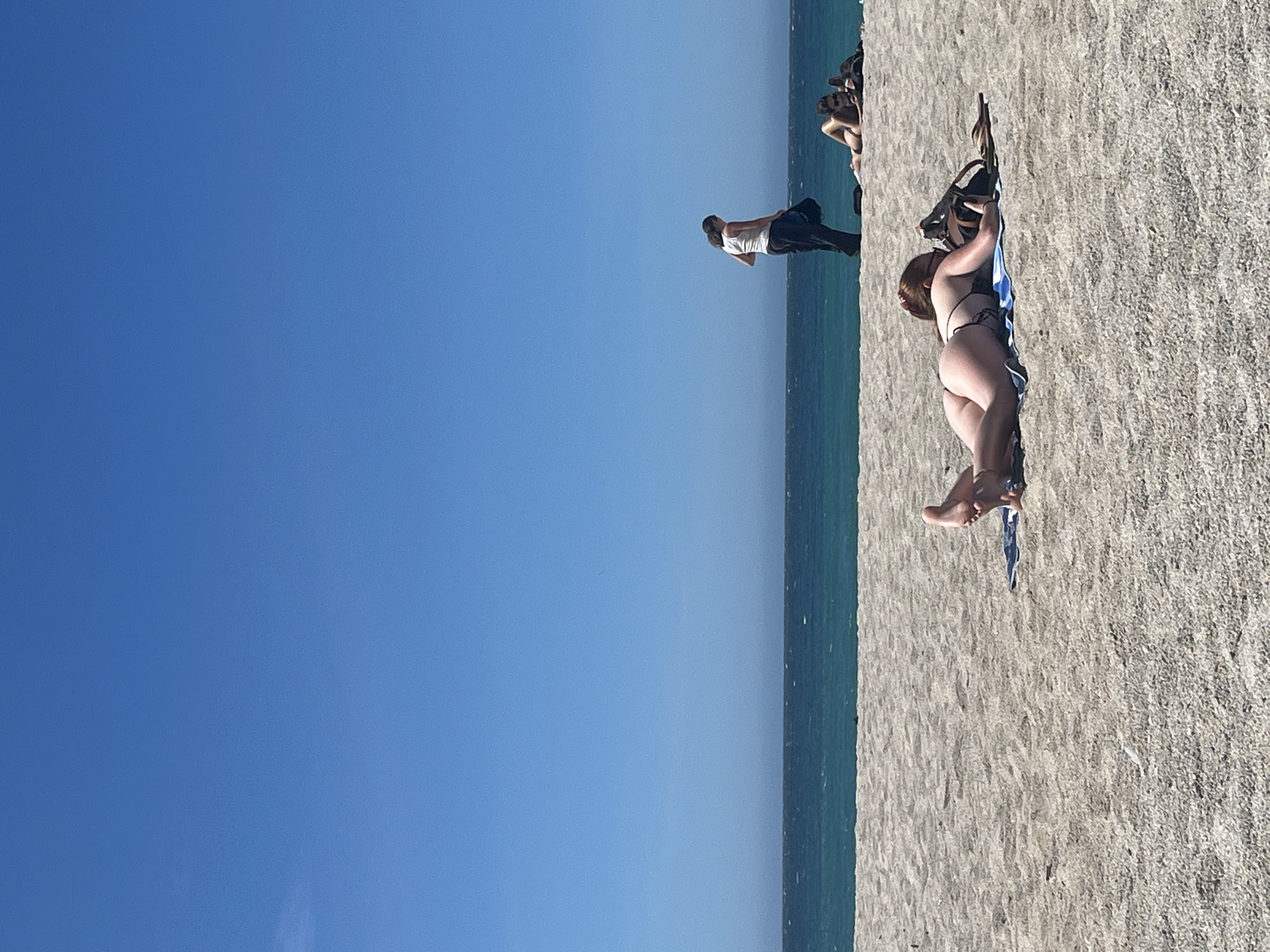 On a bright, sunny day at a beach, the foreground is dominated by a person lying face down on a towel, wearing a bikini. Their legs are bent at the knees, with their feet pointing upwards. To the right of this person, there is a bag and a pair of flip-flops. In the mid-ground, another person stands, wearing a white tank top and dark pants, holding a bag. To their right and slightly behind, two other individuals are visible lying on the sand. The background consists of a calm, blue ocean meeting a clear, bright blue sky. The scene does not depict any recognizable landmarks or features specific to the city of A Coruña, Spain.