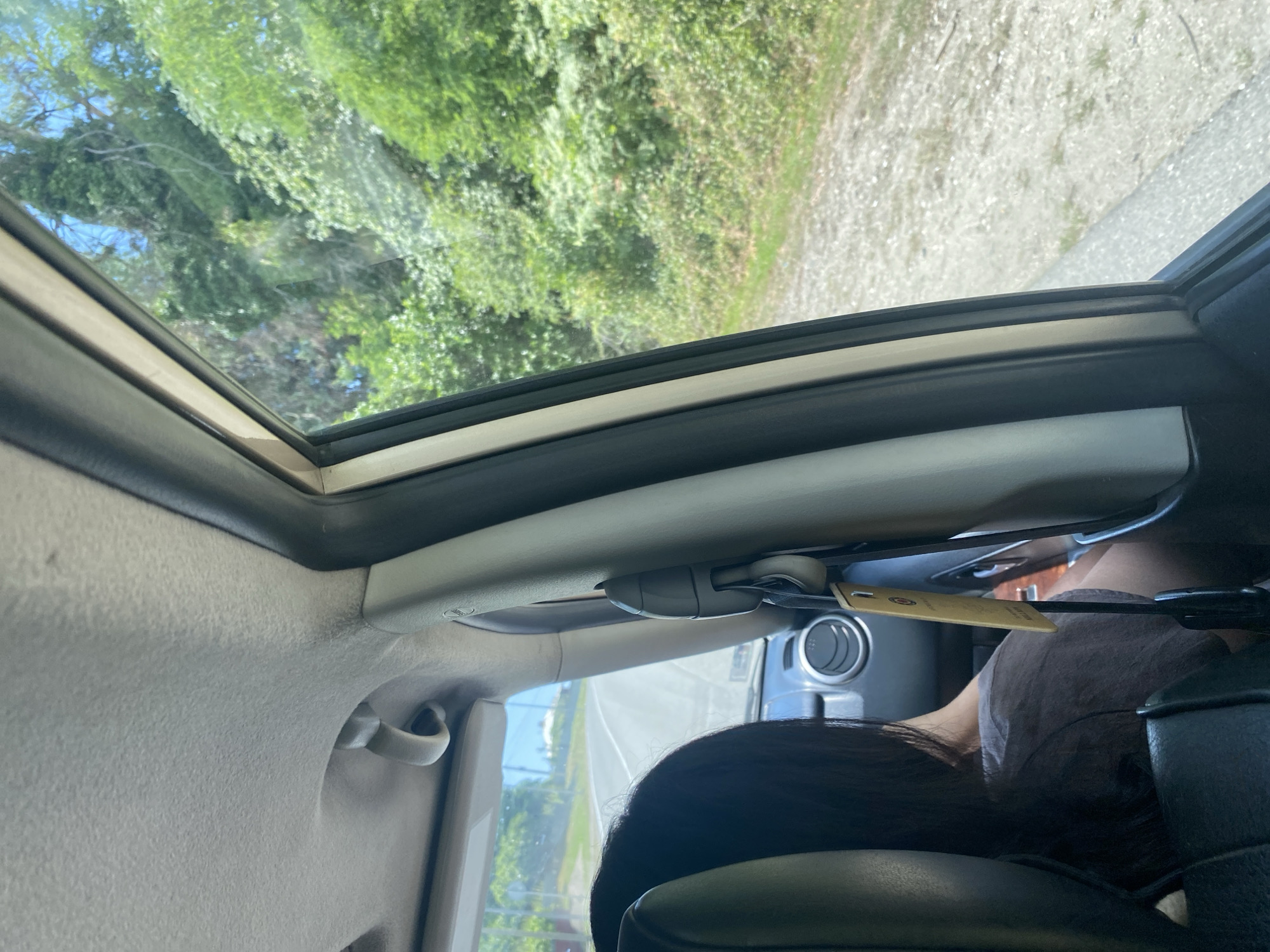 The image shows the interior of a car, looking out the passenger side window. The viewpoint is from the back seat, focusing on the headrest of the seat in front, a person's dark hair, and the car's interior trim. Outside the window, lush green trees and vegetation are visible, with a dirt road in the foreground. The sky appears bright and sunny. The scene suggests a person is traveling by car through a natural, possibly rural, environment.