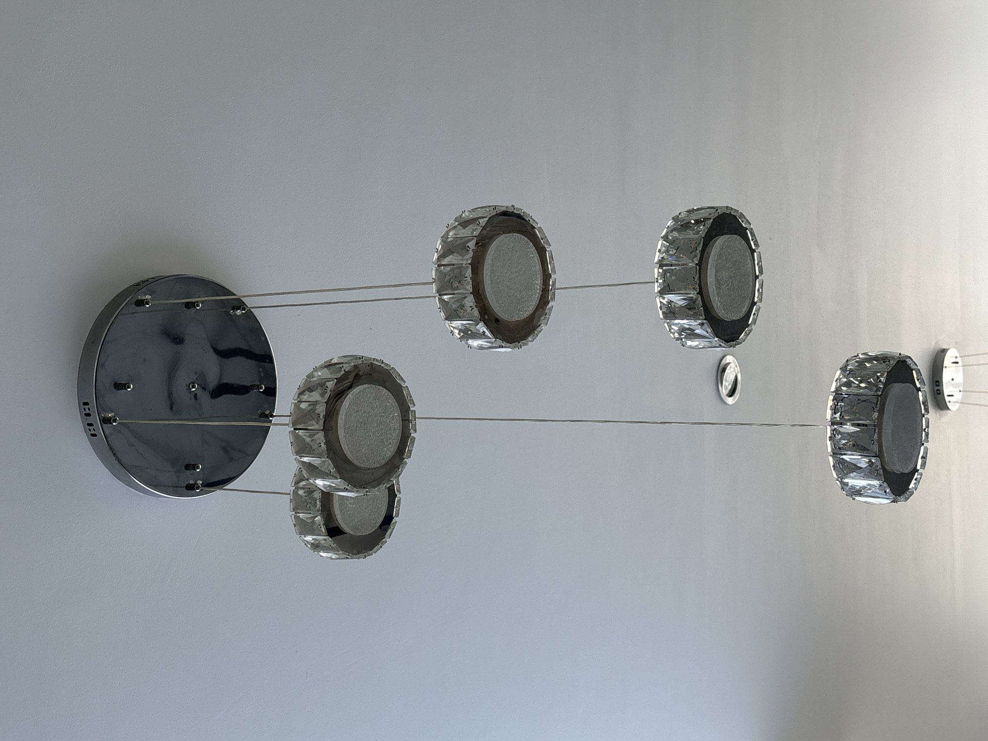This close-up, upward-angled shot captures a modern multi-pendant lighting fixture against a light grey ceiling in San Bernardino Tlaxcalancingo, Mexico. The fixture features a large, circular, chrome-plated base plate mounted flush to the ceiling on the left side of the frame. Thin, silvery wires extend from this base, suspending five main circular light elements and one smaller, simpler silver disc further to the right.
Each of the five prominent light elements is circular, characterized by a faceted crystal rim that reflects light, enclosing a frosted or textured grey circular center. These elements are arranged at varying heights and horizontal distances from the base, creating a dynamic and sculptural composition. Two elements are positioned closer to the base, another two further right, and one more is partially visible below them. A smaller, simpler silver disc is located towards the far right of the image, also suspended by a wire.
The chrome base plate is highly reflective, showing subtle distortions from the surrounding environment. On its left edge, the letters "BKB" are faintly visible, along with other small, indistinct markings. The overall design conveys a contemporary and clean aesthetic, highlighting reflective surfaces and crystal detailing. The room appears to be well-lit by ambient light, as the fixture itself is not illuminated. The background is a plain, light-colored surface, likely a ceiling or an upper portion of a wall.