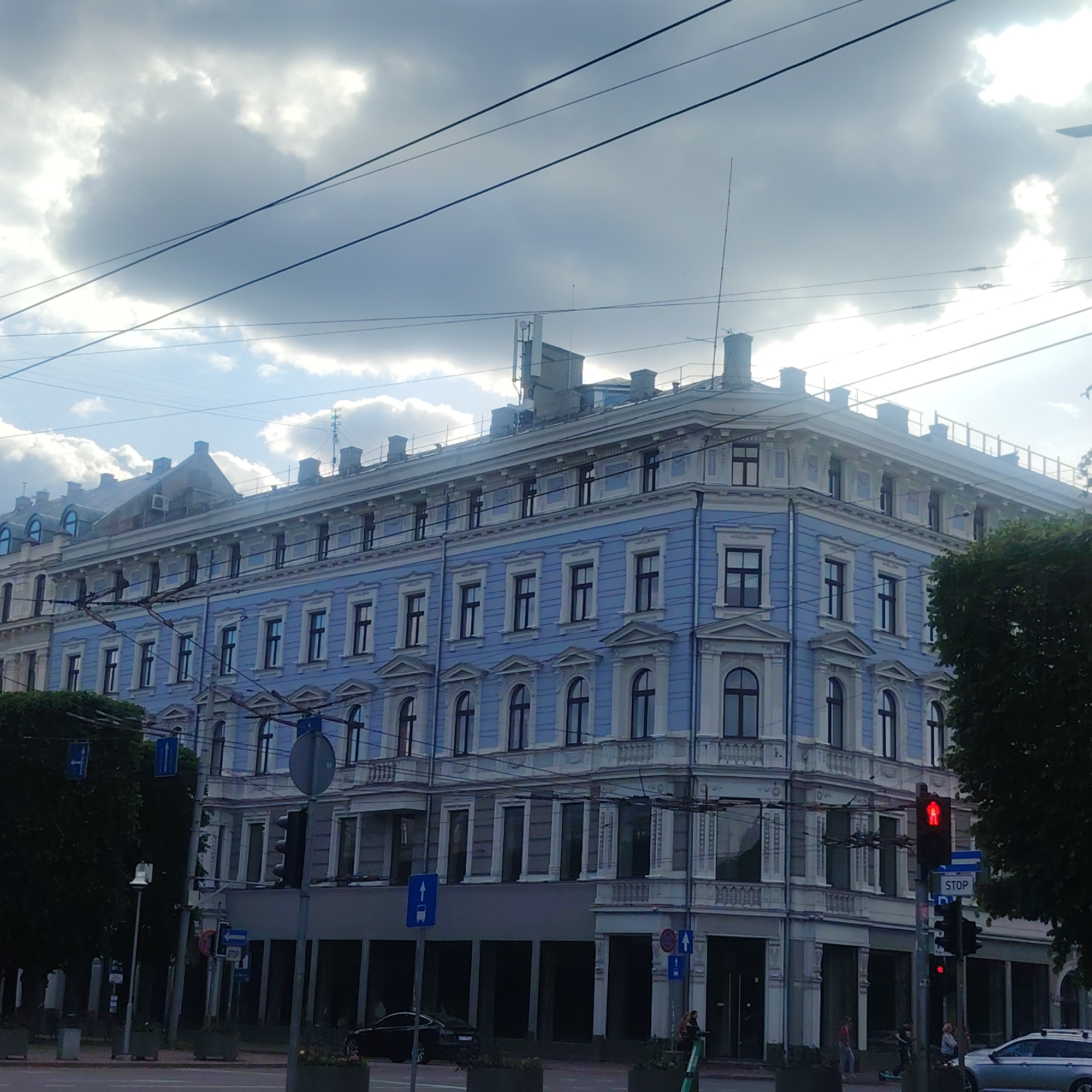 The image captures a street-level view of a multi-story building, likely in Riga, Latvia, given the distinctive architecture and overhead tram lines. The building features a light blue facade with white decorative elements around the windows and cornices. The windows are predominantly rectangular, with some upper-level windows having arched tops. The building's roofline is adorned with numerous chimneys and decorative architectural features.
Overhead, a network of thick black cables, presumably for trams or electricity, crisscrosses the frame. The sky is dramatic, with large, textured clouds partially obscuring a bright, diffused light source, suggesting either early morning or late afternoon.
In the foreground, there are traffic lights, street signs, and planters. A pedestrian signal shows a red walking figure, indicating a stop. Several dark trees flank the left side of the street, partially obscuring the lower floors of the building. On the right side, a dark green tree canopy is visible.
On the street level, there are signs of urban activity. A black car is parked on the left, and a silver car is visible on the right. People are present on the sidewalk; two individuals are standing near a scooter on the right, and others are faintly visible in the background. The overall impression is of a bustling urban environment under a dynamic sky.