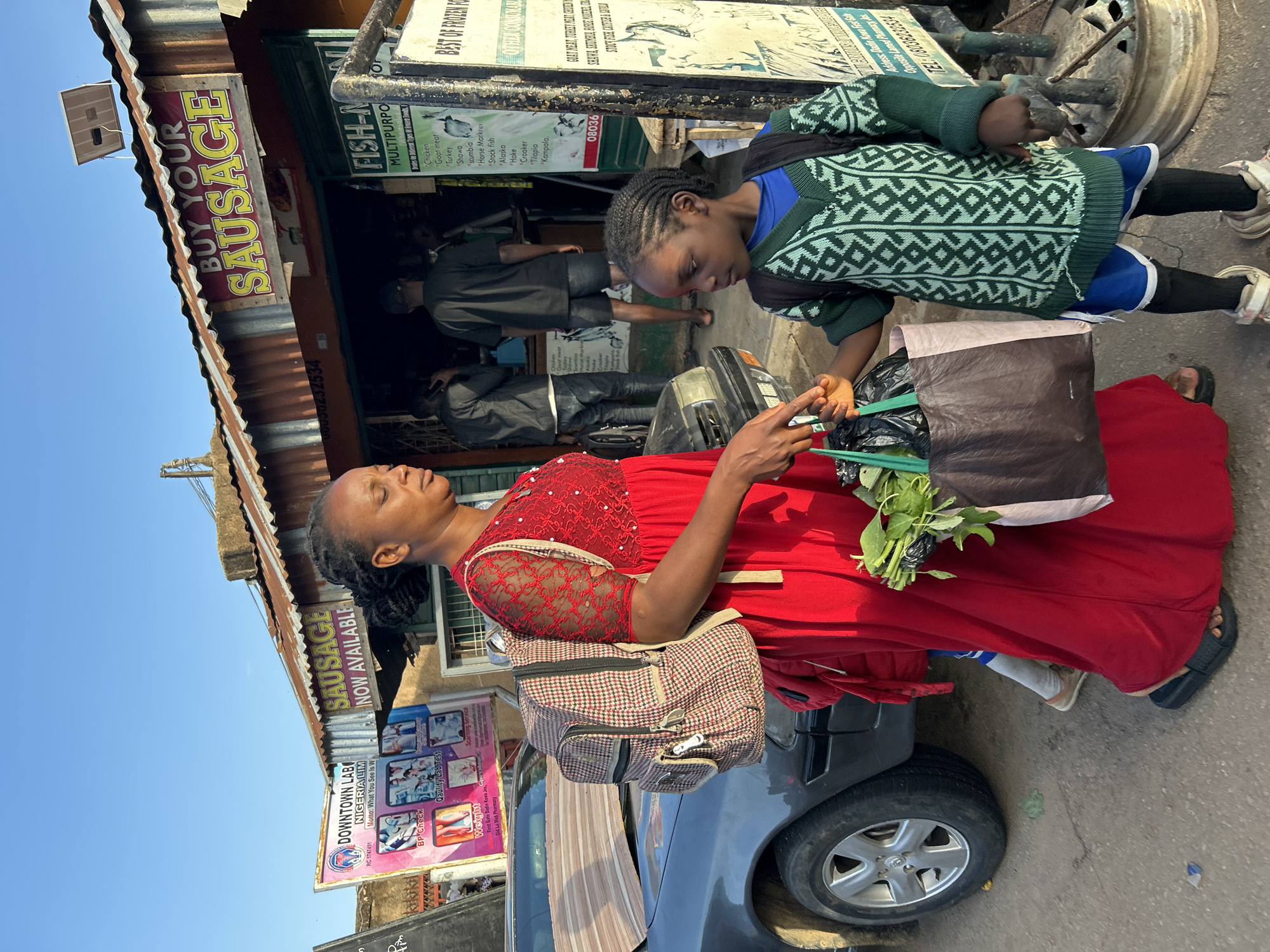 A medium shot captures a woman in a red dress standing in front of a storefront, apparently interacting with a young girl in a green sweater. The storefront, identified by a sign proclaiming "BUY YOUR SAUSAGE," is part of a business selling frozen foods including chicken, goat meat, turkey, and various fish. The signage suggests the location is in Jos, Nigeria, specifically near "First Gate Dadin Kowa," and mentions "Old La Med Pharmacy." The woman carries a brown and white plaid backpack and is holding green leafy vegetables in a black plastic bag. The girl looks toward the vegetables. The scene also includes a parked gray car with a visible tire, and signs for "DOWNTOWN LAB NIGERIA LIM" can be observed.