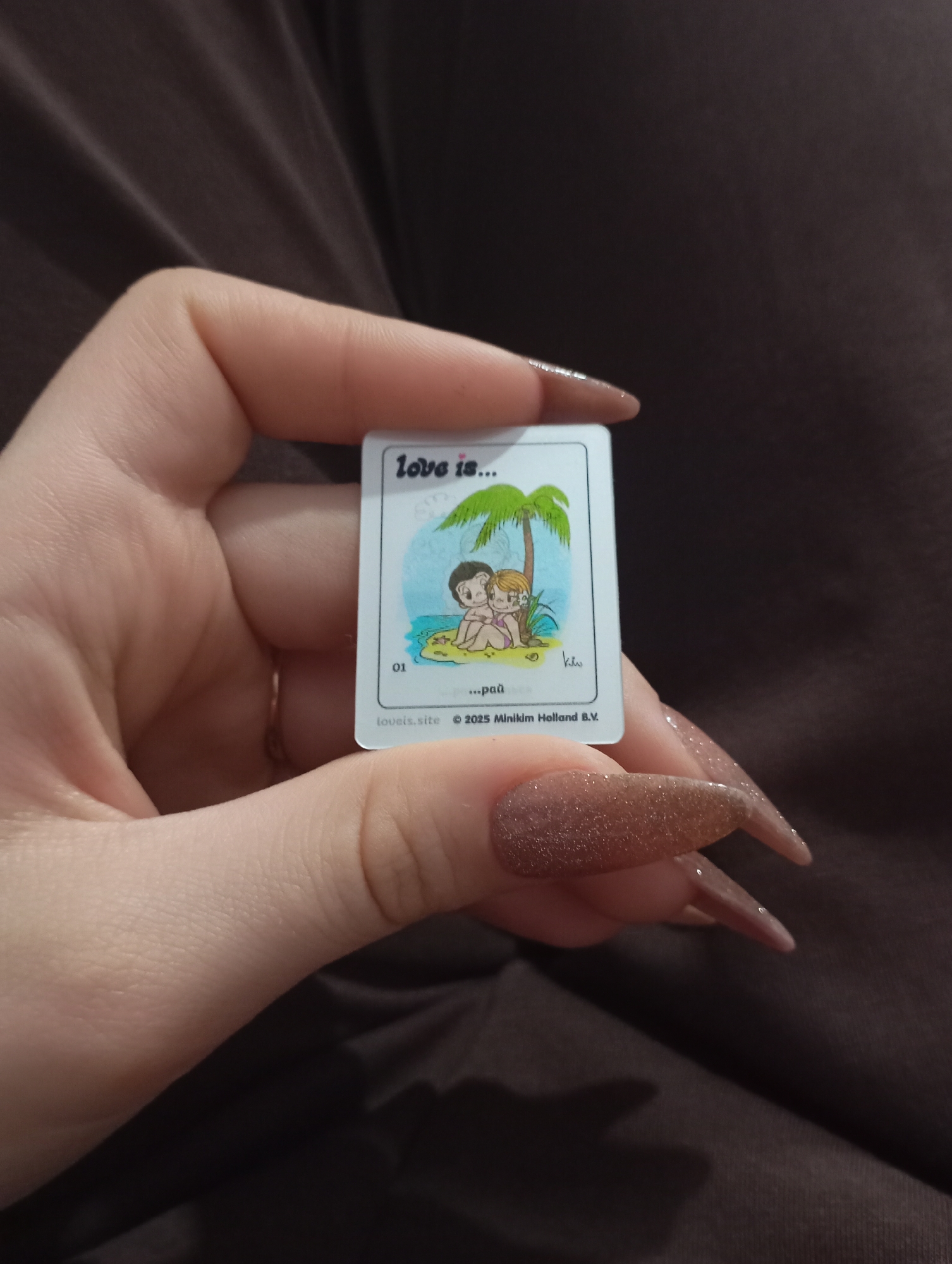 A hand holds a "Love is..." trading card. The card depicts a cartoon couple under a palm tree on a beach. Text on the card reads: "Love is...", "01", "...раū", "loveis.site", and "© 2025 Minikim Holland B.V.". The background is blurry and appears to be dark brown fabric. There is no indication of Krasnodar, Russia.