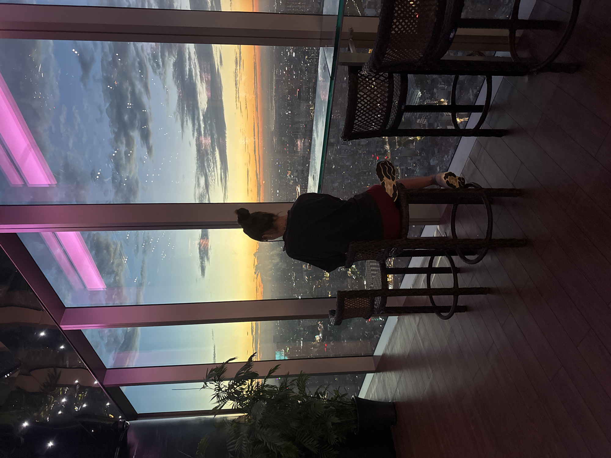 A person sits on a chair by a large window overlooking a city at sunset. The sky is a mix of blues, grays, and oranges, with clouds visible. The city below is illuminated by lights, suggesting it is dusk or early evening. The person is wearing a dark top and red bottoms, with sneakers. Their hair is pulled back in a bun. Two wicker chairs are visible, along with a wooden floor. The scene appears to be from a high vantage point, possibly a restaurant or bar, given the presence of bar stools and the view. The overall mood is peaceful and contemplative, with the person gazing out at the expansive cityscape. There is a potted plant in the foreground, adding a touch of nature to the urban setting. The pink lighting on the window frames suggests artificial illumination within the space. No visible text is present in the image.