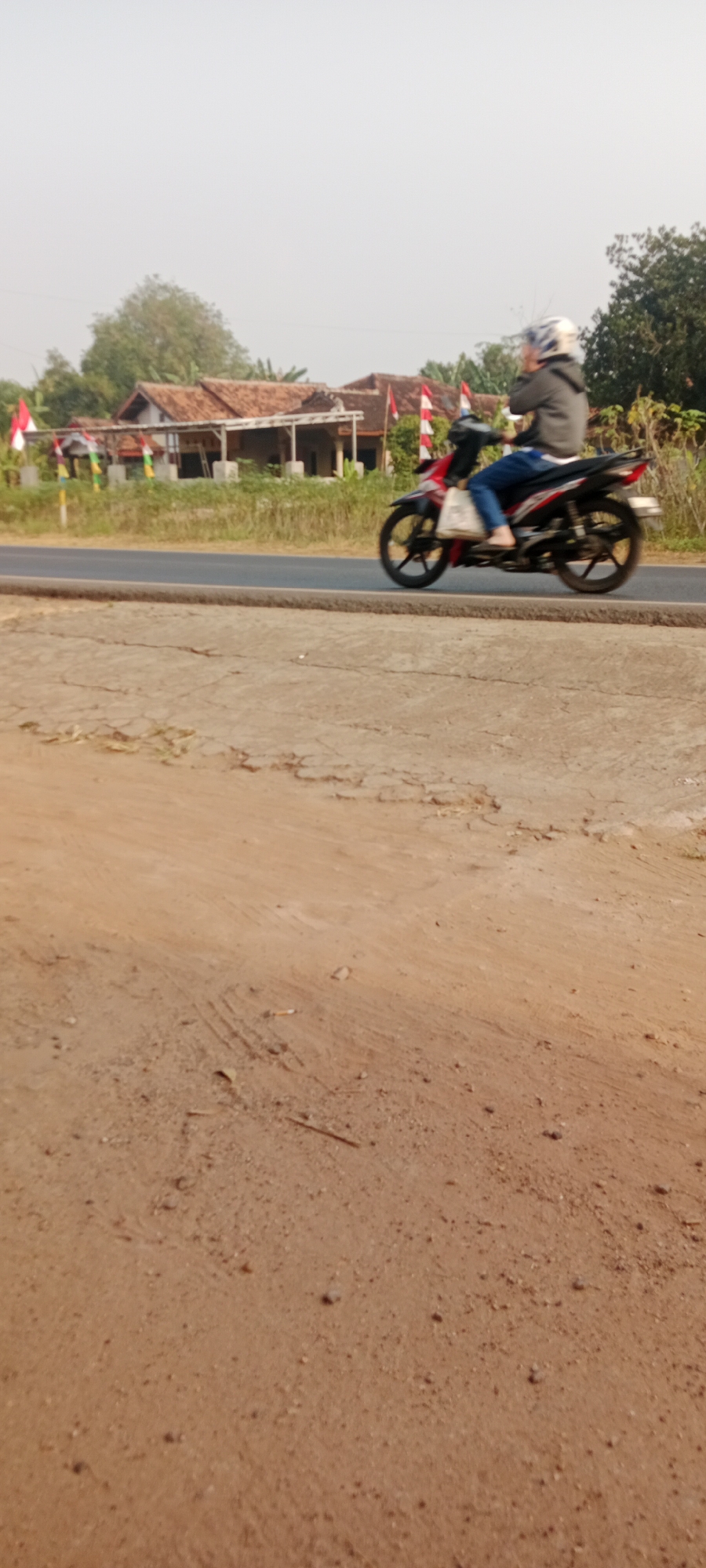 A person rides a red and black motorcycle down a paved road. The rider is wearing a grey jacket and blue jeans, and a white helmet with a black stripe. The person is carrying a white bag on the left side of the motorcycle. In the background, there are several houses with red tile roofs and Indonesian flags displayed. The road is bordered by dry grass and trees. The foreground shows a dirt path with some small rocks and dried vegetation. The sky is overcast and hazy. The scene appears to be located in a rural area of Gondang Rejo, Indonesia.