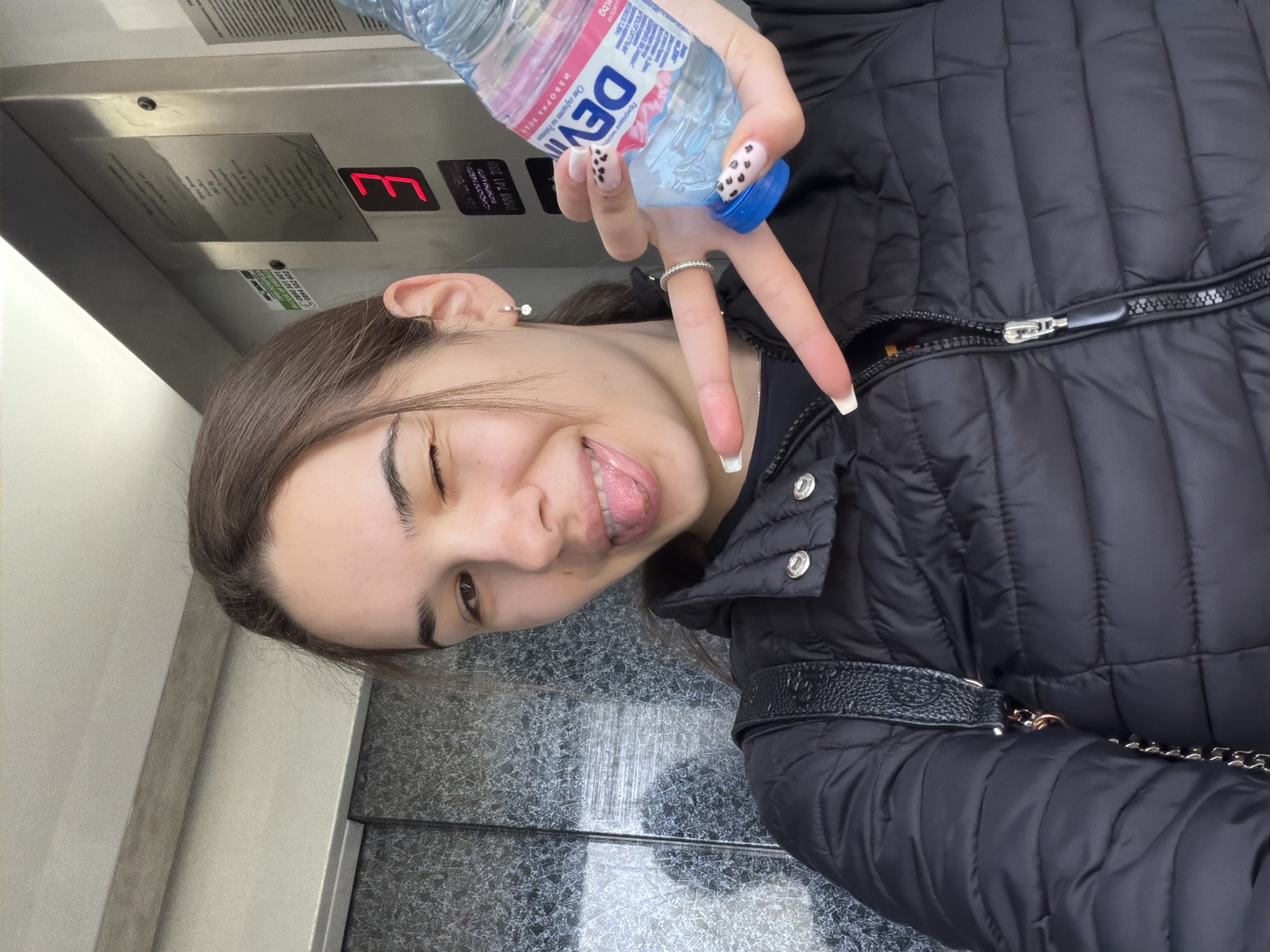 Here's a forensic description of the image:
The image appears to be a selfie taken in what seems to be an elevator. A young woman is the main subject. She is wearing a black, padded jacket and a ring. She is holding a bottle of "Devin" brand water, with a label in Bulgarian. She is making a peace sign with her fingers. The background consists of the elevator's interior, including the floor indicator showing "E", and metal walls.
There's not enough information in the picture to definitively determine if this is in the city of Sofia, Bulgaria. The water bottle is a brand sold in Bulgaria, however.