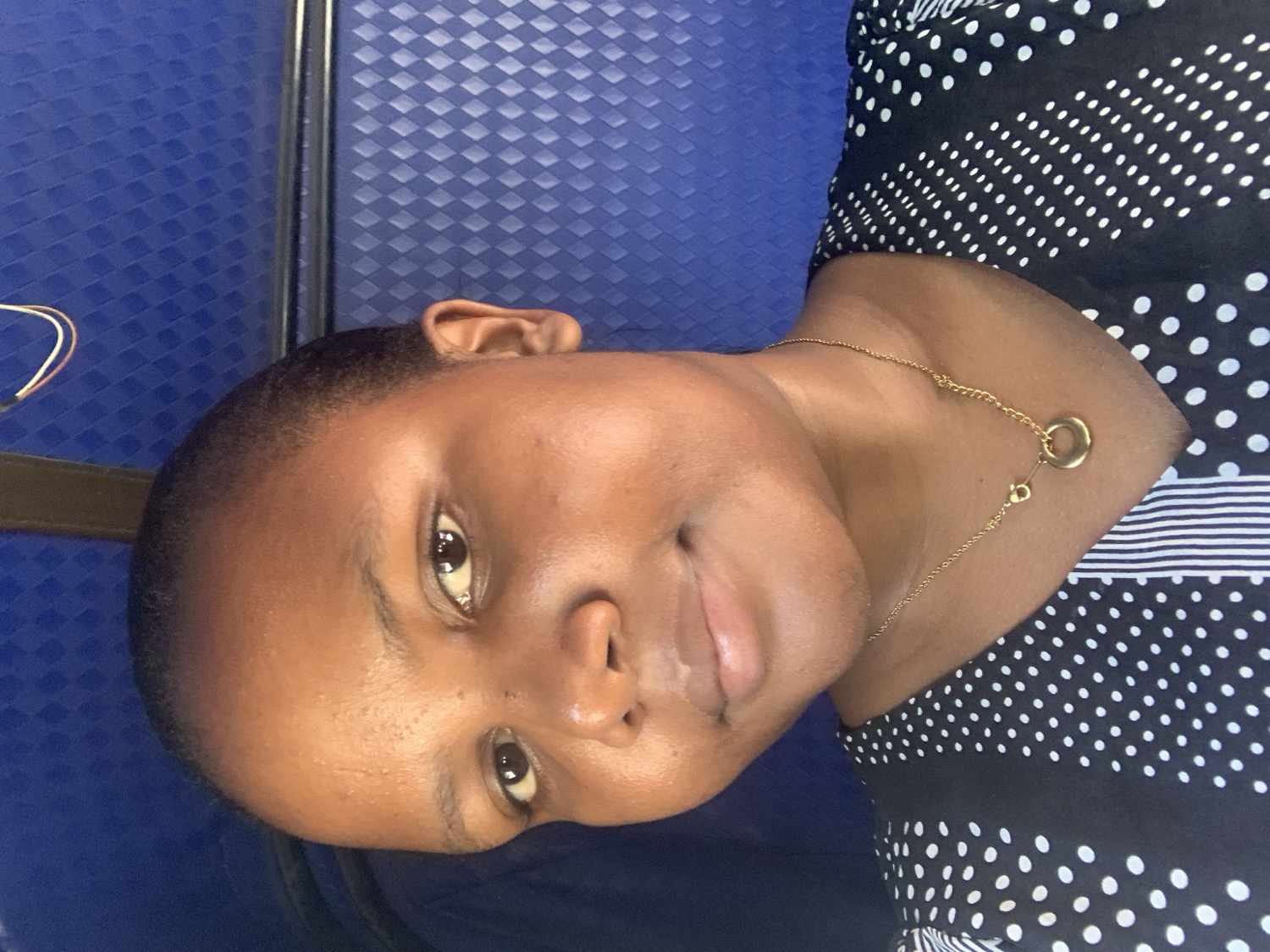 The image features a young Black woman captured in a close-up, chest-up shot. The original image is rotated 90 degrees clockwise, showing her head tilted to the right. When viewed upright, she is looking directly at the camera with a gentle, friendly smile on her lips, her dark eyes open wide. Her hair is cut very short.
She is wearing a black top adorned with white polka dots of varying sizes, and a fine gold chain necklace with a small, circular, open-ring pendant hangs around her neck, fastened with a visible clasp.
The background primarily consists of a textured, dark blue surface, possibly a seat or wall, patterned with raised square or diamond shapes. A thin, dark, vertical strip, possibly a metal rail or seam, runs along the left side of the background. To the far left, a glimpse of a light-colored wire and a dark strap can be seen.
The activity taking place is a posed portrait. The lighting is soft and even, suggesting an indoor or shaded environment, possibly within a vehicle or waiting area in Dar es-Salaam, Tanzania. Her expression conveys calmness and approachability. No visible text is present in the image.
