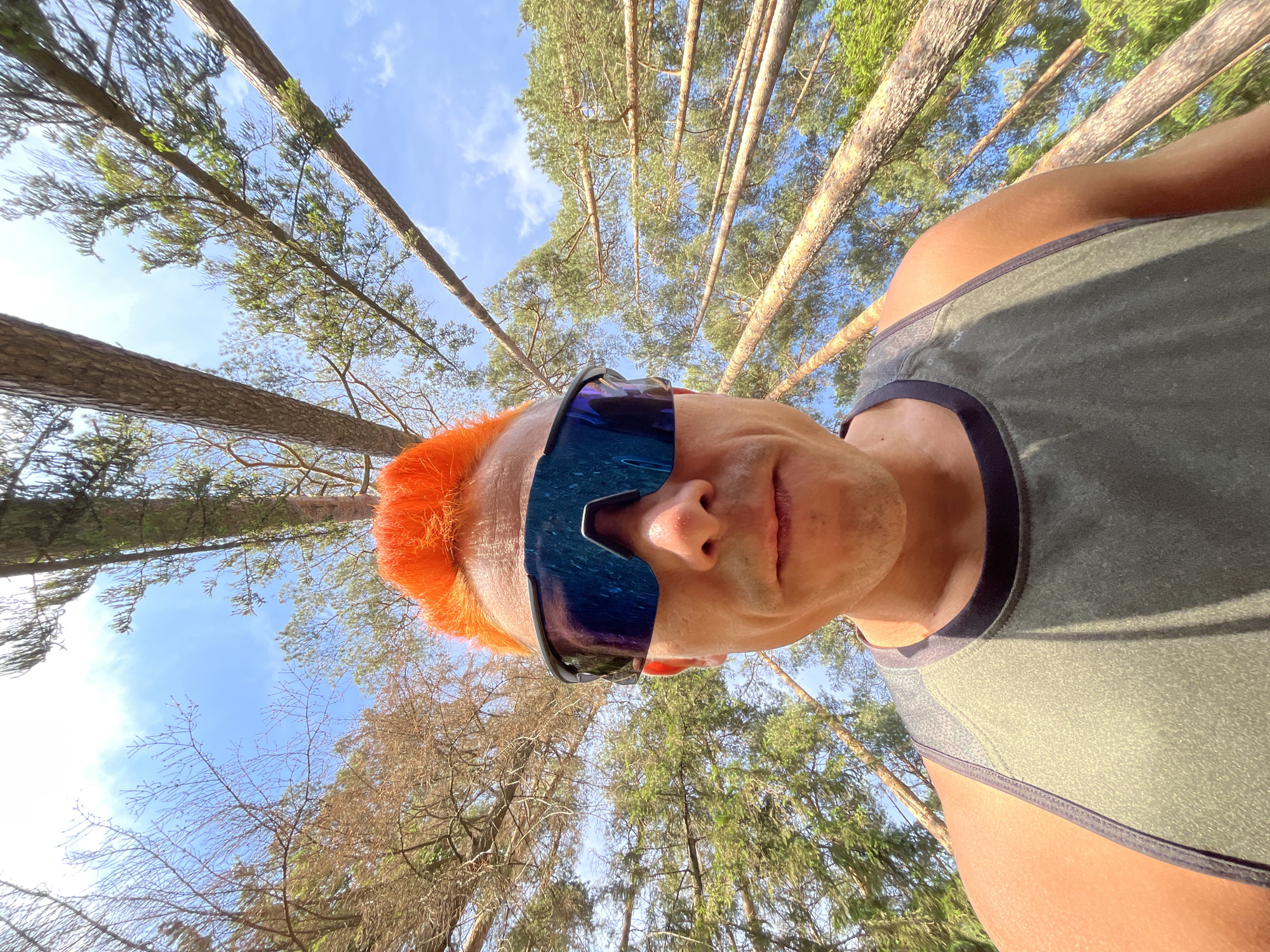 A low-angle, close-up outdoor selfie shot features a man with orange hair wearing blue mirrored sunglasses and a gray tank top. The background reveals a dense forest with tall trees stretching upward toward the sky. The location appears to be a wooded area in Chaikovsky, Russia, based on the type of trees. The sky is visible through the tree canopy, exhibiting scattered clouds.