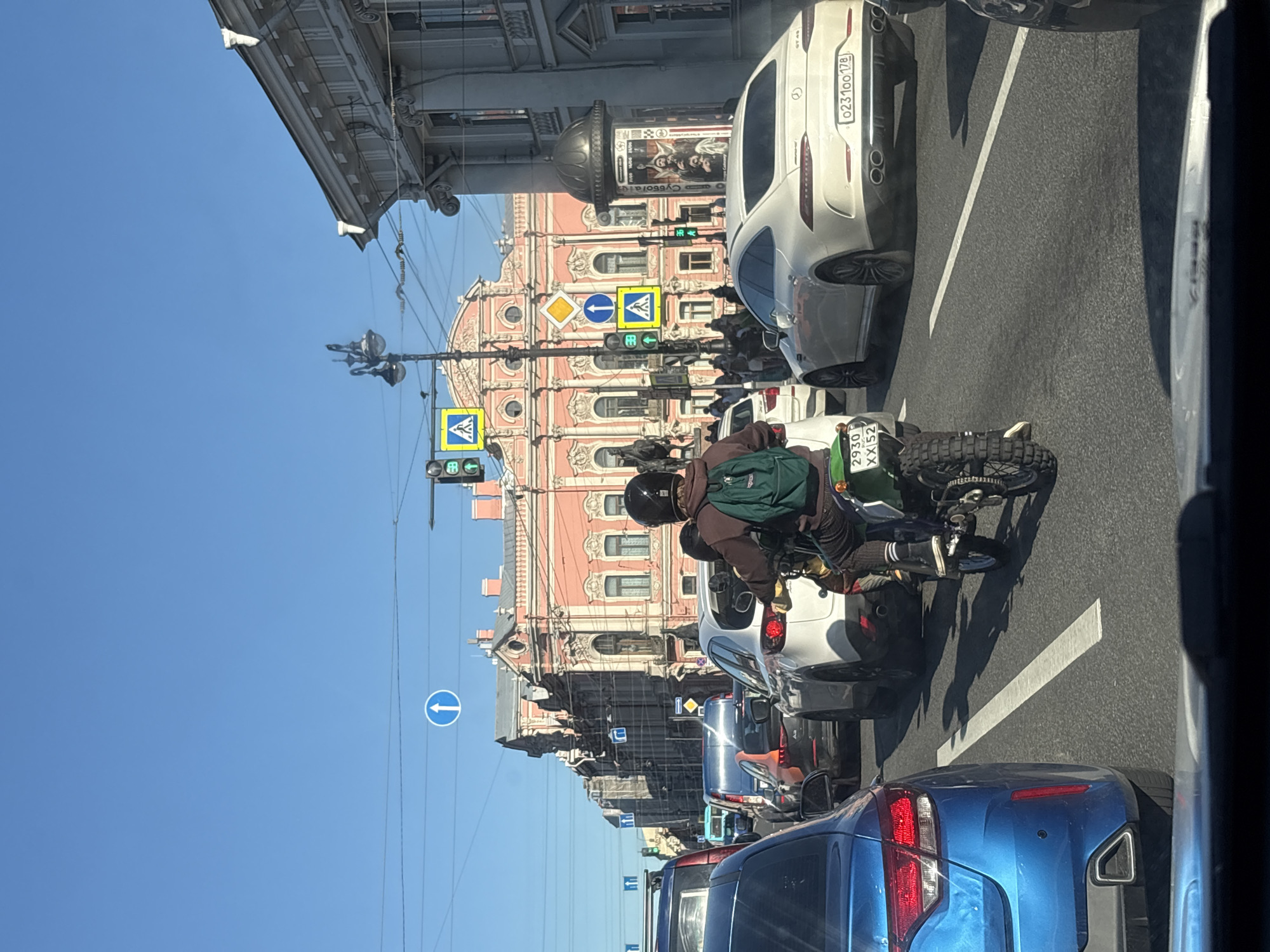 The image depicts a daytime scene on a busy urban street in Saint Petersburg, Russia, observed from the interior of a vehicle looking forward. The sky is clear and blue, and the lighting is bright, casting shadows on the asphalt.
In the immediate foreground, the rear section of a blue car is partially visible on the left. Centrally positioned, a person on a green dirt bike or enduro motorcycle is seen from behind. The rider wears a dark helmet, a brown jacket, dark pants, and a green backpack. The motorcycle has prominent knobby tires and displays a Russian license plate "2930 XX 52".
Ahead in traffic, a white Mercedes-AMG GT 4-door Coupe is on the right side of the frame, showing a Russian license plate "0231OO 178", confirming the Saint Petersburg region. To the left of the motorcycle, a white Mazda CX-5 SUV is partially visible. Further back on the left, an orange sedan or SUV is discernible amidst the stationary or slow-moving traffic.
The background is dominated by architectural elements. A large, ornate, multi-story building with a salmon-pink facade and classical architectural features, including columns and decorative window surrounds, stands prominently in the center. To the far right, a lighter grey building with classical cornices and pilasters is visible. Overhead, numerous utility lines crisscross the blue sky.
Various street signs are present, including a blue circular sign with a white upward arrow (mandatory direction), multiple yellow square signs with black pedestrian symbols (pedestrian crossing), and active traffic lights. A tall, ornate black lamppost is situated on the left side of the road. On the right, adjacent to the white Mercedes, a cylindrical advertising column features a poster with religious iconography and Cyrillic text. The road surface shows solid white lane markings.