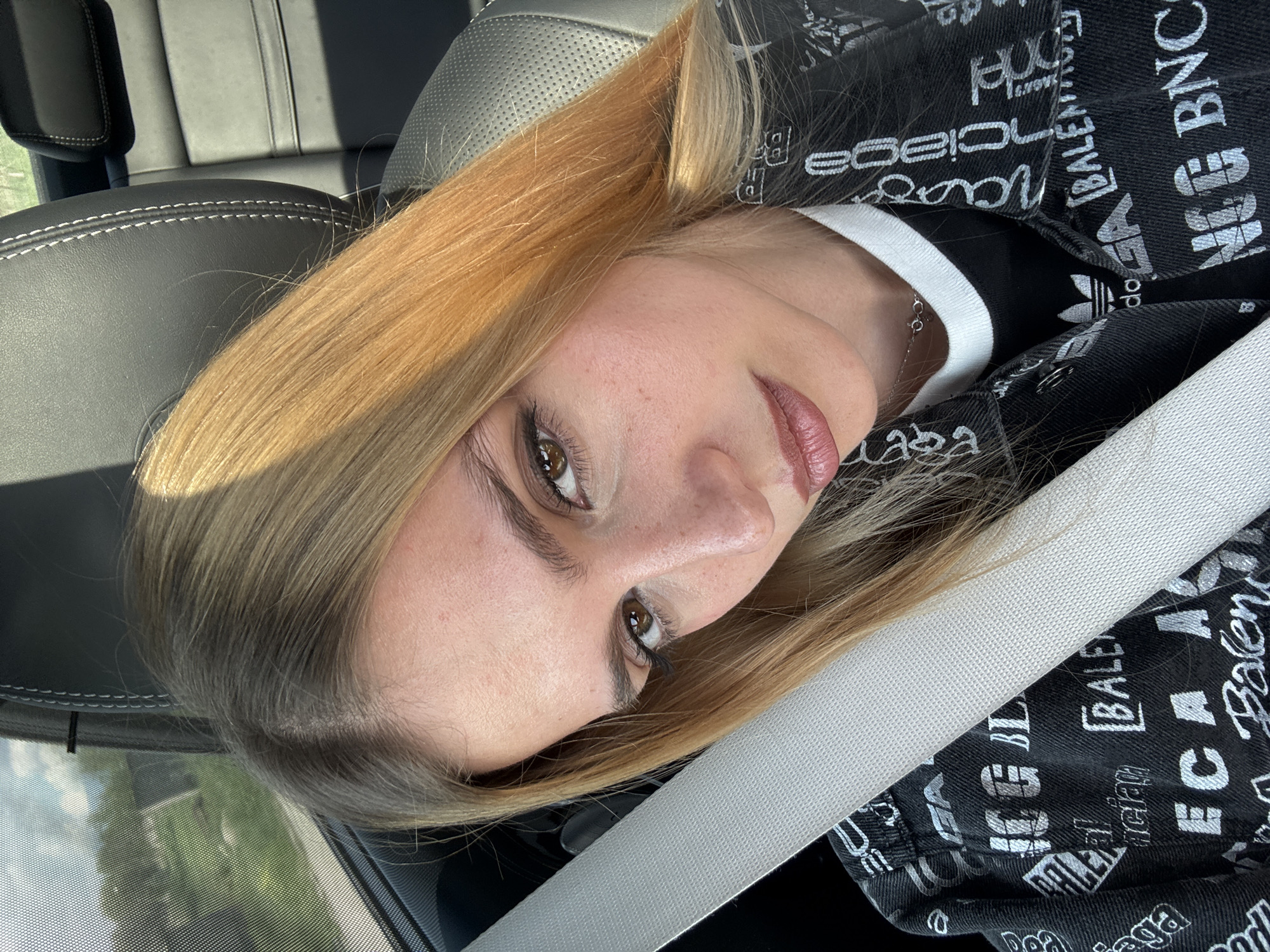 A photograph presents a woman with blonde hair and brown eyes, wearing a black jacket patterned with white lettering. She is seated in a car, as evidenced by the seat belt across her body and the car's interior. The location is not identifiable.