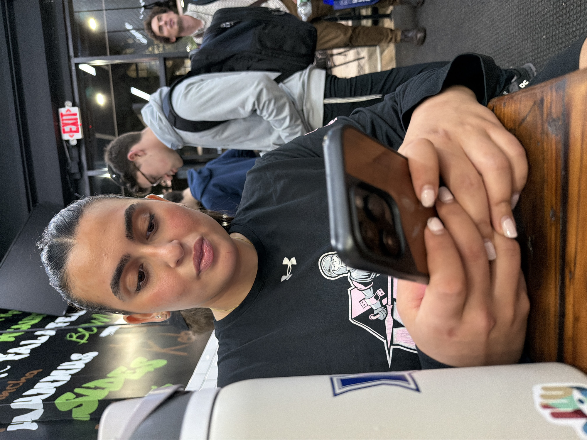 A photograph taken in New York City, United States, shows a woman seated at a wooden table, holding a smartphone. The woman is wearing a black t-shirt with an athletic logo. In the background, other people are visible, including two men standing. An "EXIT" sign is visible on a wall.