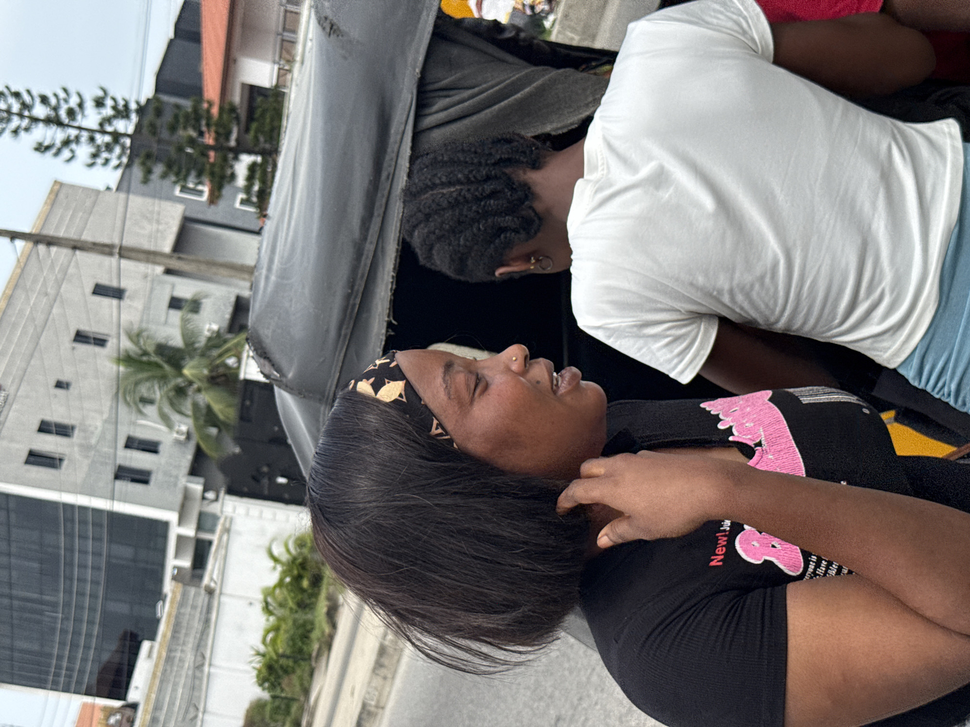 The image shows two individuals inside a vehicle. One woman, with dark hair and a patterned headband, is visible from the shoulders up, wearing a black shirt with a pink design. She is turned towards the left of the frame, with her hand near her neck. Another individual, wearing a white shirt and blue shorts, is seen from the back, leaning forward within the vehicle. The vehicle appears to be a three-wheeled transport.
The background reveals an urban environment with buildings of varying heights, including a modern structure with numerous windows. Palm trees and other greenery are interspersed among the buildings. The scene appears to be in Ado, Nigeria.