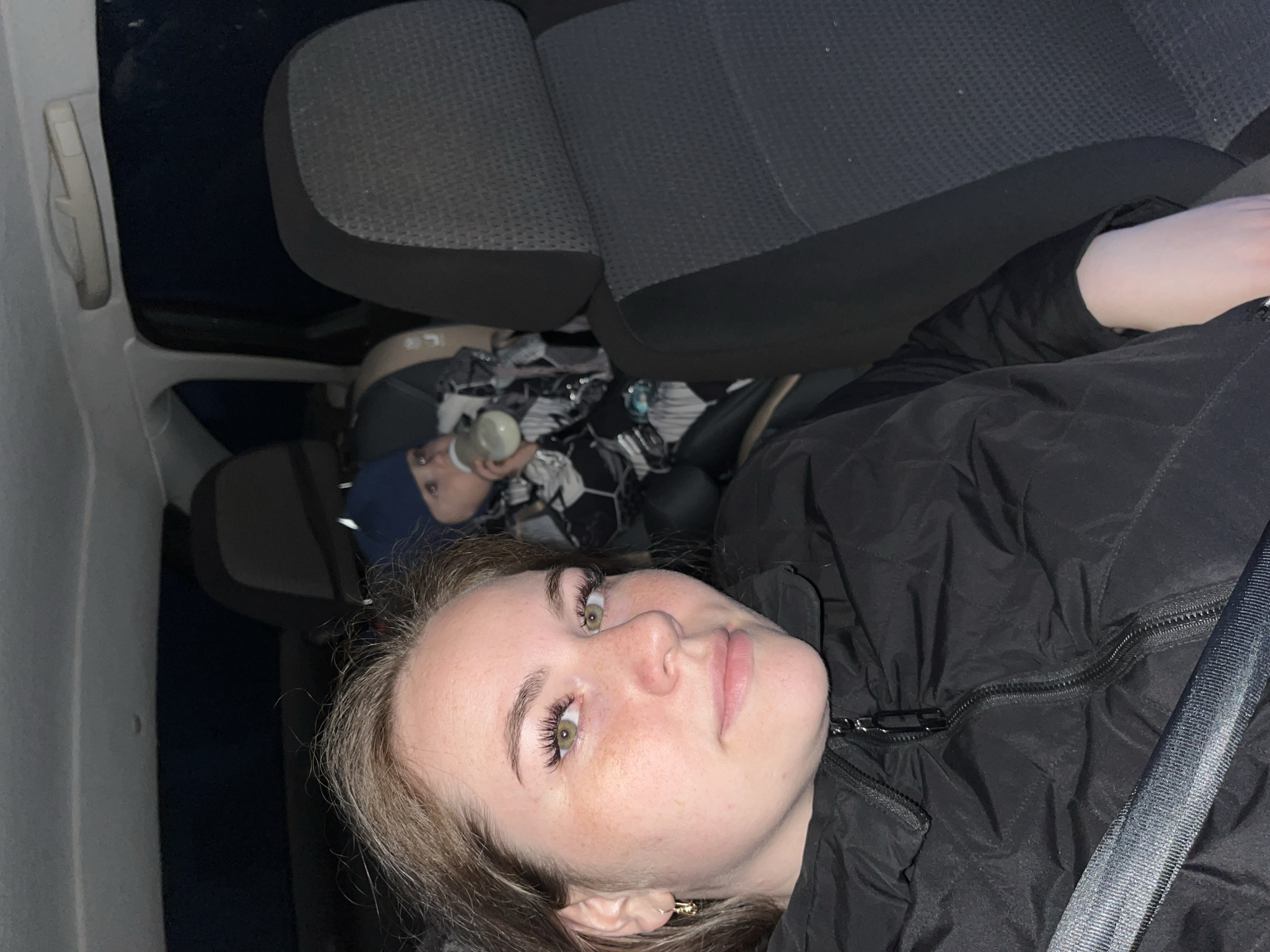 The image appears to be taken inside a car, possibly in transit. A young woman with light skin, light brown hair, and green eyes is in the foreground, wearing a black jacket and a seatbelt. A baby, dressed in a blue hat and patterned clothing, is visible in the background, holding a bottle. The seats of the car are dark gray. The location is provided as Chaikovsky, Russia, though this cannot be verified from the image alone.
