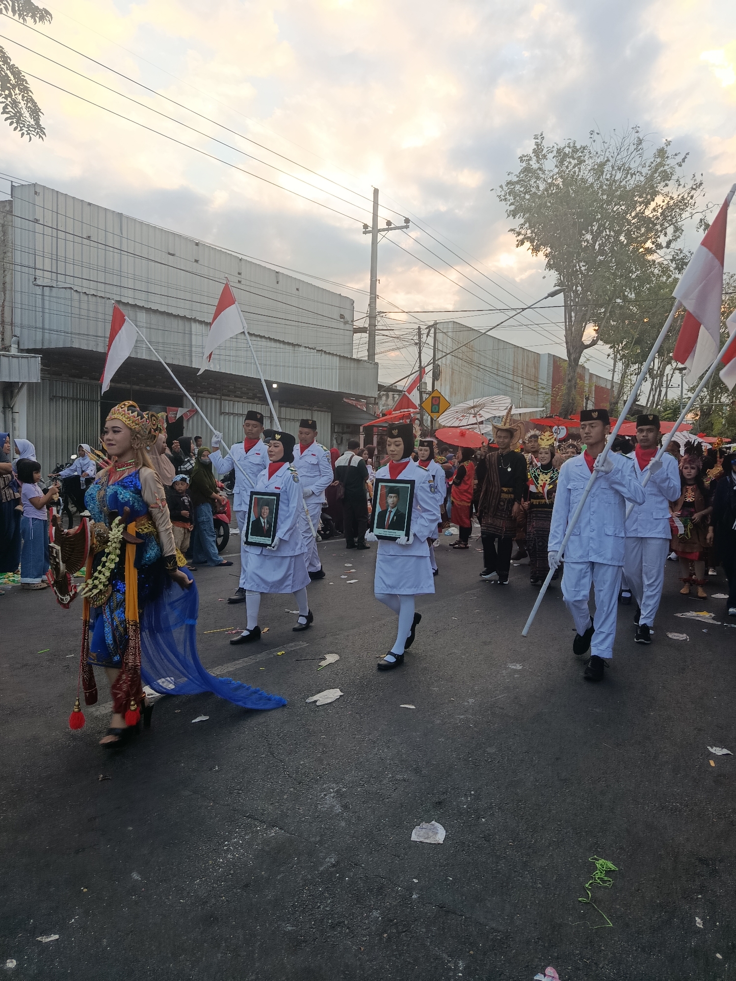 In Ardirejo, Indonesia, a parade is taking place on a paved street under a cloudy sky at what appears to be dusk. The foreground features a person in traditional Javanese attire, complete with an ornate headdress, elaborate jewelry, and a blue and gold costume. To the right, a group of individuals in white uniforms, likely students or cadets, are marching in formation. Some of them are holding Indonesian flags and framed portraits of what appear to be past national leaders. In the background, buildings line the street, and other parade participants and spectators are visible, some riding motorcycles. The atmosphere suggests a celebratory or commemorative event.