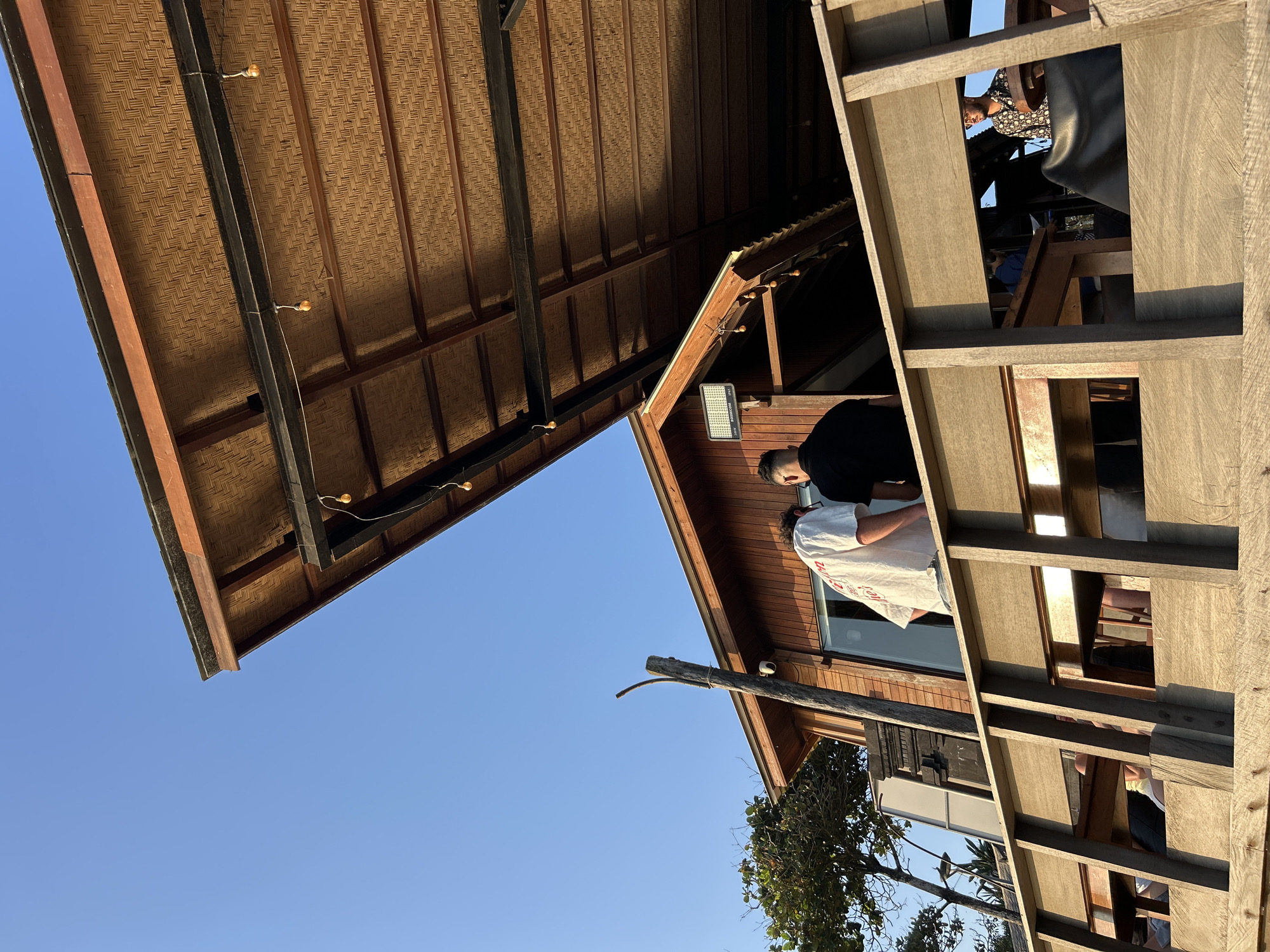 Two individuals stand on a wooden balcony, silhouetted against a clear blue sky. One person wears a white shirt with red lettering, while the other is dressed in black. They are positioned near a wooden building with a thatched roof, characteristic of beachside establishments in North Kuta, Indonesia. The building features large windows and an outdoor seating area with tables and chairs, some occupied by other patrons. String lights hang from the underside of the roof, adding to the relaxed, tropical ambiance. A weathered wooden post stands near the building, and lush green foliage is visible to the left, suggesting a coastal setting. The overall scene captures a moment of leisure and social gathering at a waterfront location.