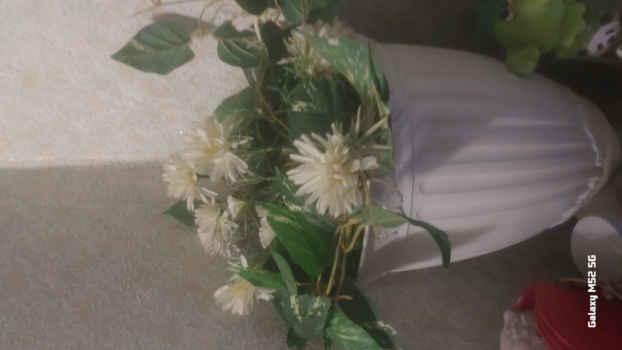 The image depicts a still life arrangement of artificial flowers and foliage within a white, fluted vase. The flowers are creamy white and have a daisy-like appearance with multiple thin petals. Green leaves, some with variegation, are intertwined with the flowers. The vase is positioned in what appears to be an indoor setting, with textured walls in the background. A small, green, frog-shaped figurine is visible to the right of the vase. The bottom left corner of the image contains the text "Galaxy M52 5G" in white lettering, indicating the device used to capture the photograph. The scene does not provide any specific details that would allow for precise localization within the city of Netishyn, Ukraine; it is a generic indoor composition.