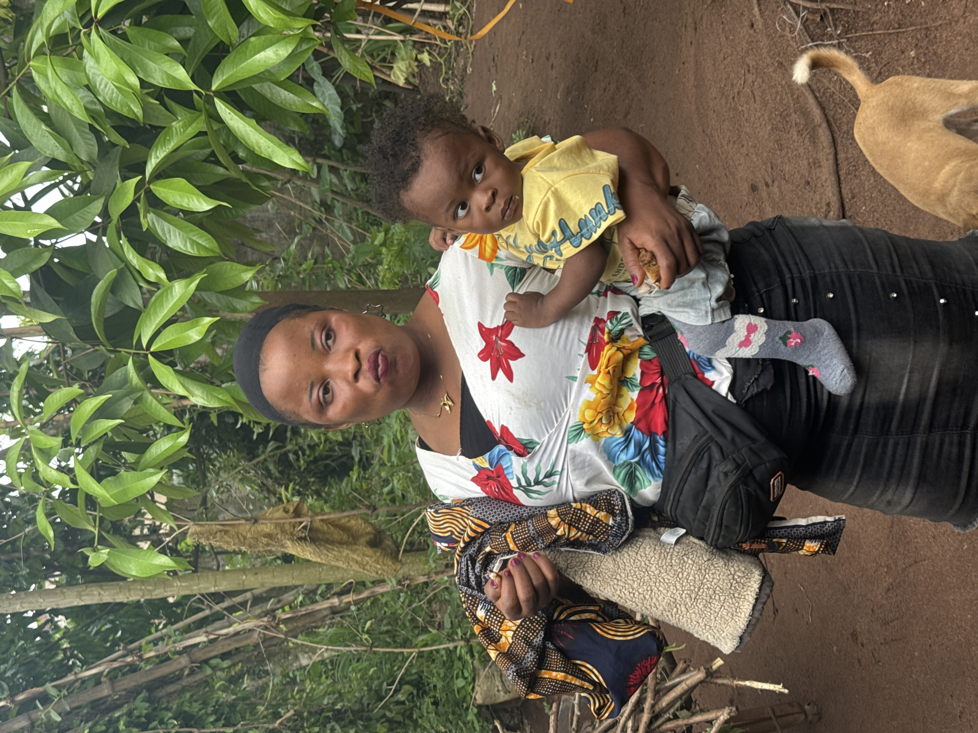 A woman stands in an outdoor area with trees and a dirt ground. The woman is holding a small child. She wears a black headband, a floral white blouse, a black shirt underneath, a patterned fabric around her waist, black pants, and a small black bag. The child wears a yellow shirt, light blue shorts, and gray socks with floral patterns. The woman appears to be looking at the camera with a direct gaze. The child is also looking at the camera. Part of a dog is visible in the upper right corner. The location appears to be a yard or garden, potentially in the city of Aboh, Nigeria.