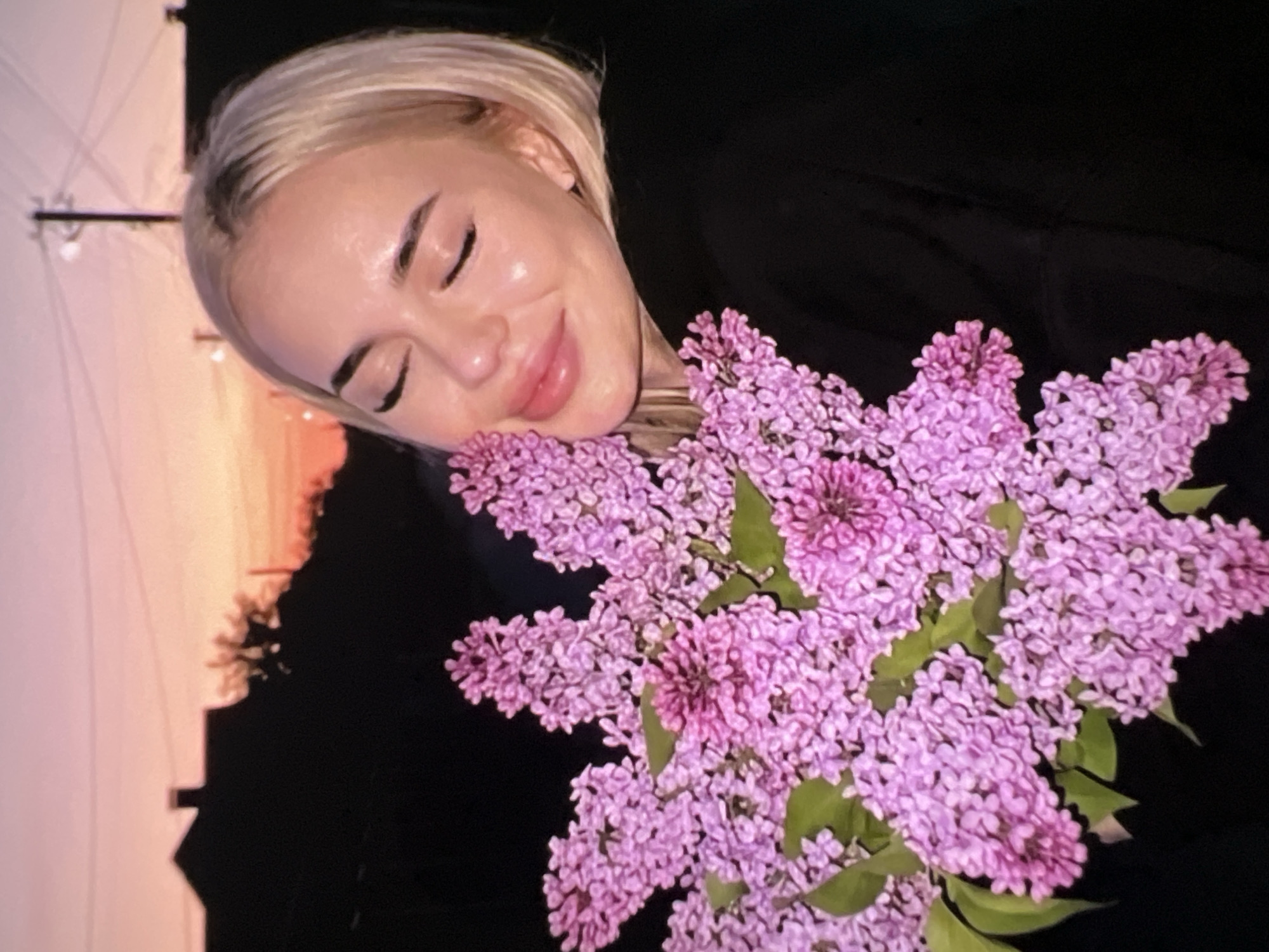 A rotated portrait-style image captures a young woman with her eyes closed, gently smiling, and holding a substantial bouquet of lilac flowers close to her face. The woman has fair skin and light blonde hair, with well-defined eyebrows and pinkish lips. She appears peaceful and content.
The prominent bouquet consists of numerous clusters of vibrant purple lilac blossoms, displaying a range of shades from light lavender to a deeper violet, with some green leaves interspersed throughout. She is dressed in a dark garment, largely obscured by the flowers and the dark background.
The background on the left side of the image features a soft, warm gradient sky, indicative of either sunrise or sunset, transitioning from pale pink and orange at the top to a richer orange and red further down. Silhouetted against this sky are a utility pole with wires, and dark, indistinct shapes of structures and trees, suggesting an outdoor scene or a view through a window. The right side of the image is largely dark, providing a striking contrast to the woman's bright face and the colorful flowers.
The overall atmosphere of the image is serene and joyful, conveying a moment of quiet appreciation for nature's beauty. There is no visible text in the image.