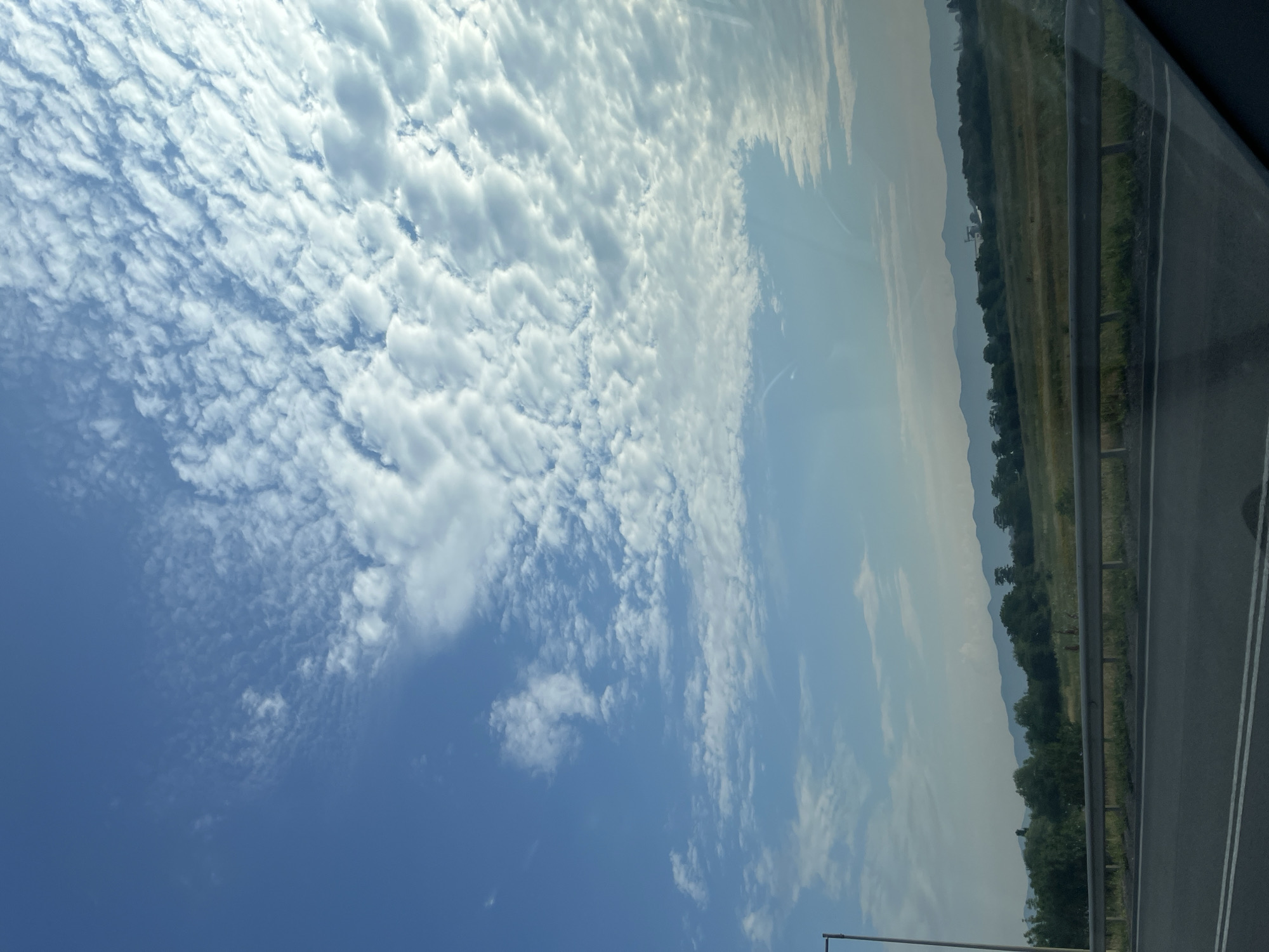 The image captures a scenic view of the sky and landscape, possibly from a moving vehicle, as suggested by the perspective and road in the foreground. The sky is a prominent feature, filled with a vast expanse of blue with scattered, fluffy white clouds that form an interesting, textured pattern across the upper portion of the frame. These clouds appear to be mid-level cumulus clouds, with some areas showing a more broken, ripple-like formation.
Below the sky, a distant landscape unfolds. A range of rolling hills or mountains can be seen in the far background, shrouded in a light haze that gives them a soft, blue-gray hue. In the middle ground, there is a strip of green vegetation, including trees and grassy fields. A metal guardrail separates the foreground from the middle ground, suggesting the presence of a road or highway.
In the immediate foreground, a portion of a paved road with white lane markings is visible. The angle of the road suggests the photograph was taken from a vehicle driving on it. A tall, slender object, possibly a lamppost, is visible on the far left side of the image, further indicating an urban or roadside environment. The overall lighting suggests it is daytime, likely morning or late afternoon, given the soft quality of the light and the colors of the sky and landscape. There are no people or specific actions visible in the scene. The environment appears to be natural and somewhat rural, with a hint of infrastructure from the road and lamppost.