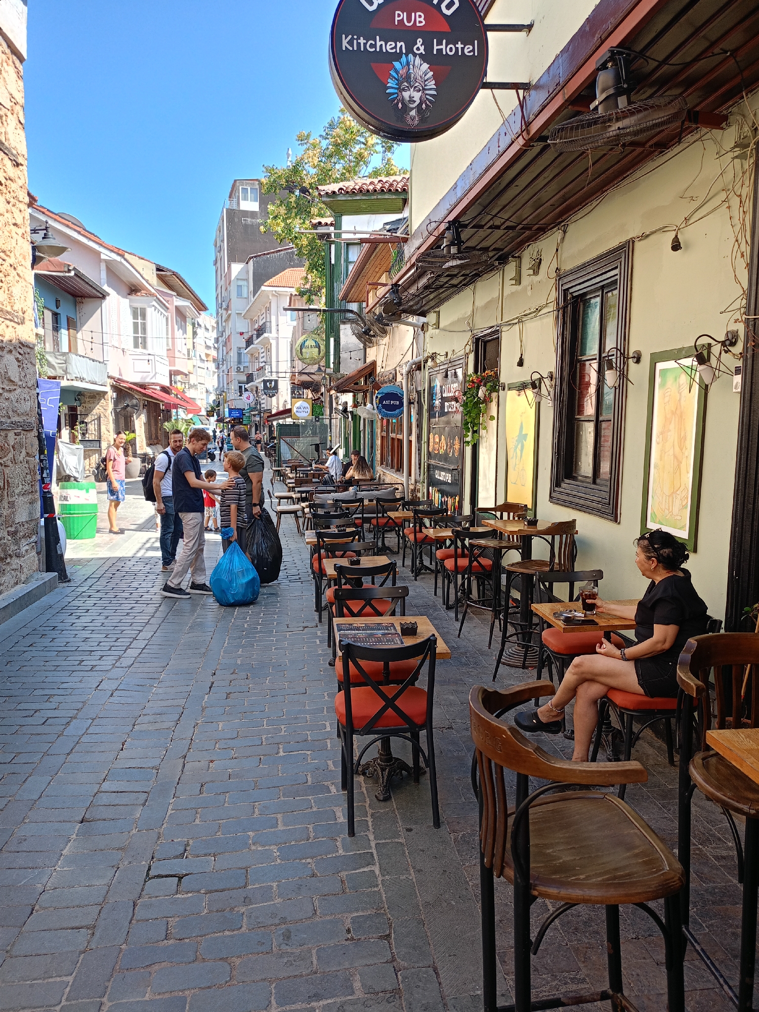 The image captures a street scene in Antalya, Turkey, likely in the Kaleiçi (Old Town) district, characterized by its narrow cobblestone streets and historic architecture. The perspective is from ground level, looking down a pedestrian walkway lined with businesses.
On the right side of the street, an outdoor seating area for a pub or restaurant is set up with numerous tables and chairs. The tables are small and round or square, mostly with orange-cushioned chairs. A woman with dark hair, wearing a black top and shorts, is seated at one of these tables, facing away from the camera and looking towards the building. She has a drink in her hand. The building facade is painted a light yellow-green, with dark-framed windows and decorative lighting fixtures. Above the pub entrance, a circular sign reads "PUB Kitchen & Hotel" with an image of a woman in Native American headdress. Fans are mounted on the overhang of the building.
On the left side of the street, a group of people are walking or standing. Four men and a boy are gathered in the middle of the walkway. One man in a dark blue shirt is speaking to a boy in a striped shirt. The boy is holding a large blue plastic bag. Another man in a white t-shirt and backpack is looking on. A woman in a pink top and shorts stands further back, near a green cylindrical bin. The buildings on this side of the street are older, with textured stone walls on the far left and painted facades with balconies further down. A narrow alleyway is visible between the buildings on the left.
The cobblestone pavement is the dominant ground texture, with visible wear and some cracks. The sky above is clear and blue, indicating a sunny day. The overall atmosphere suggests a typical lively street in a tourist area.