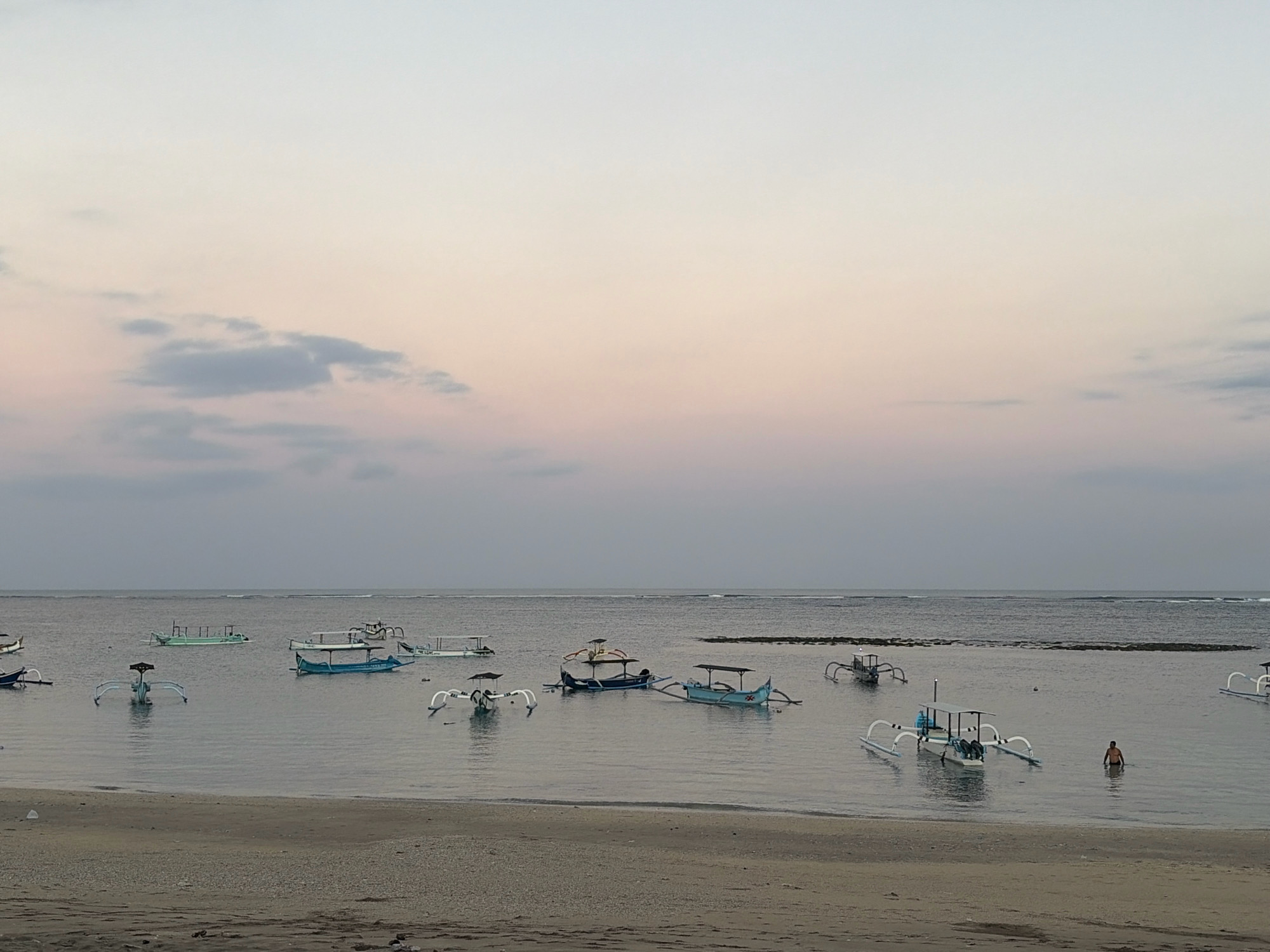 The image depicts a serene beach scene in Kuta, Indonesia, during the soft light of either dawn or dusk. The sky is a gradient of pale blue and pink, with a few wispy clouds adding texture. The calm sea is a muted gray, reflecting the sky.
Several traditional Balinese outrigger boats, known as "jukung," are anchored in the shallow water. These boats are characterized by their long, slender hulls and outriggers on either side, which provide stability. Some of the jukungs are empty, while others appear to have small canopies providing shade. The boats are varied in color, with a prominent blue one in the mid-ground and a distinctive white and blue one with a prominent dark superstructure in the foreground.
On the sandy shore in the foreground, there is a subtle texture of sand and small pebbles. The beach itself appears to be a gentle slope leading into the water.
In the water, towards the right side of the image, a lone individual, appearing to be a person, is wading. They are positioned near some of the boats, partially submerged in the water. The person's posture suggests they might be cooling off or preparing to enter a boat. The overall atmosphere is one of tranquility and a quiet moment before or after a day's activities.