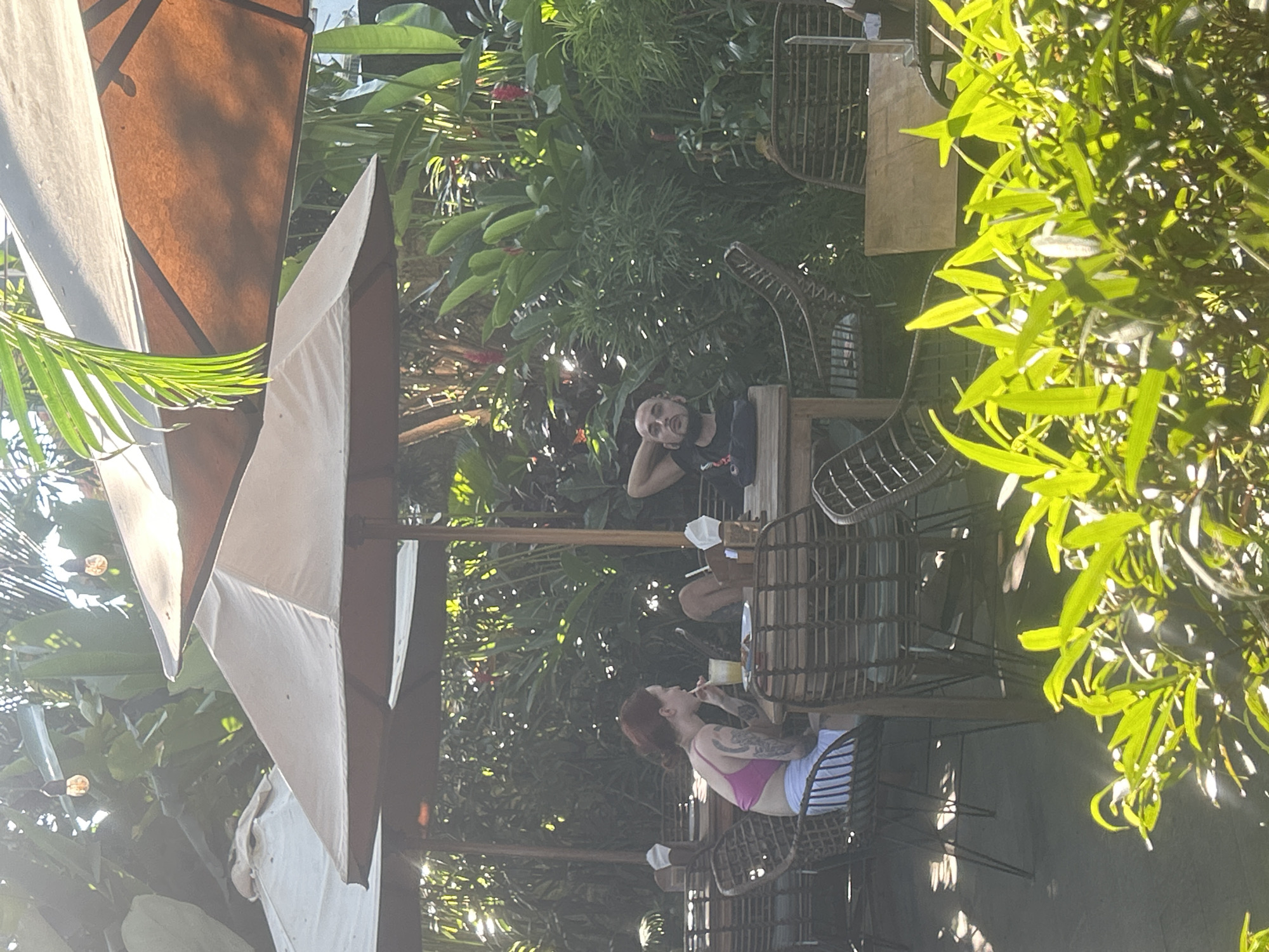 In North Kuta, Indonesia, two individuals are seated at tables under outdoor umbrellas, surrounded by lush tropical foliage. The person on the left, with red hair styled in a bun, wears a pink top and white shorts, and is actively drinking from a glass with a straw. They are seated at a wicker chair at a wooden table laden with plates. To the right, a man with a shaved head and beard is reclined in a wicker chair at another wooden table. He wears a dark t-shirt and rests his head on his hand, looking towards the camera. The setting appears to be a restaurant or cafe, with the natural light filtering through the palm fronds and umbrellas. The tables are spaced apart, suggesting an outdoor dining area.