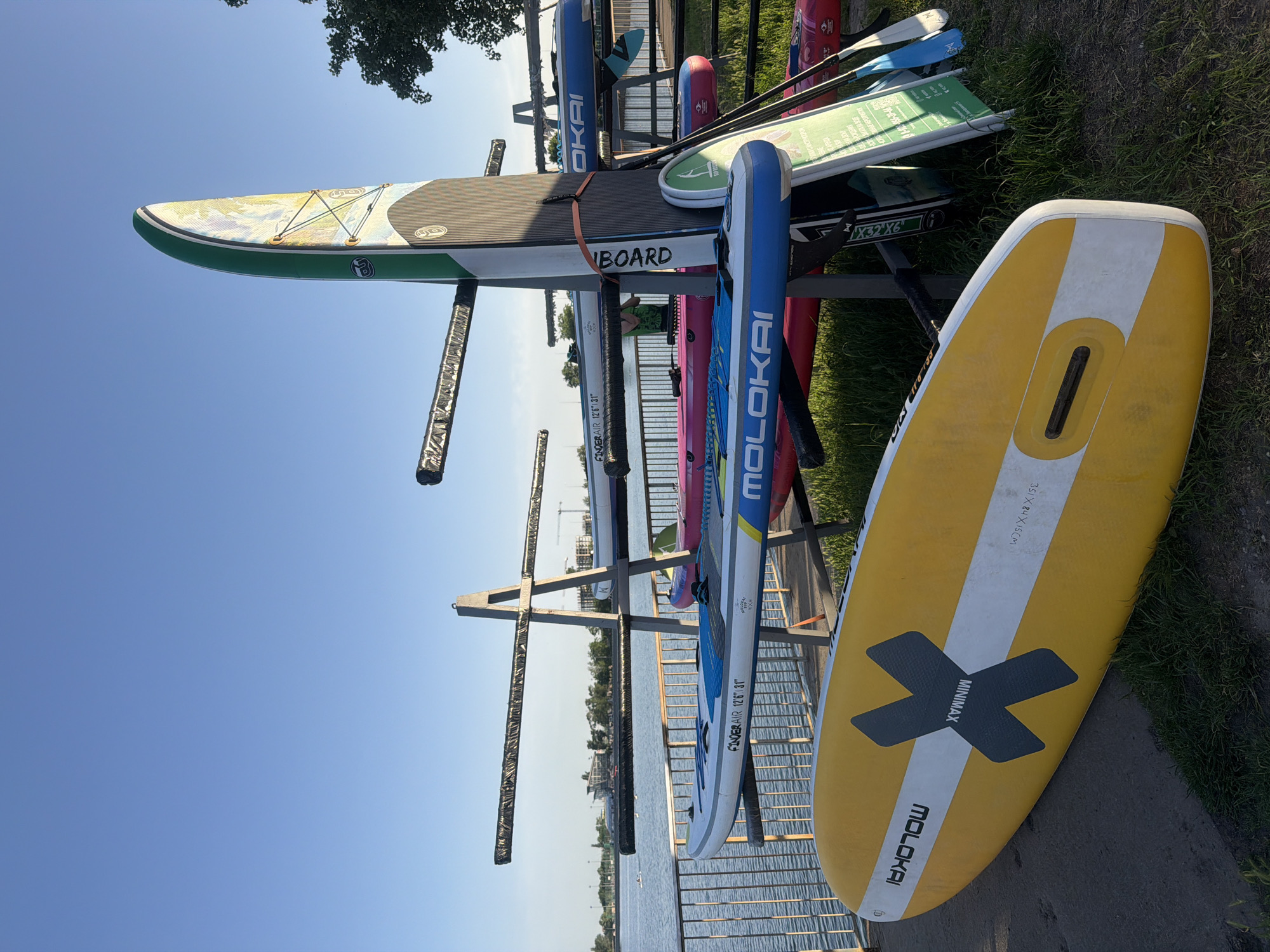 The image captures a collection of paddleboards stored on a metal rack near the waterfront in Rostov-on-Don, Russia. The rack is situated on a patch of grass alongside a paved walkway, with a metal railing separating the area from the water. The sky is clear and blue, indicating a sunny day.
Several paddleboards are visible on the rack. A yellow Molokai Minimax board with a black X graphic is propped up against the side of the rack. Various other boards are in the rack, including a green and black board, blue Molokai boards, and pink boards. Oars are also stored on the rack.
The background shows the water, with glimpses of the distant cityscape and trees. The overall scene suggests a recreational area near a body of water.