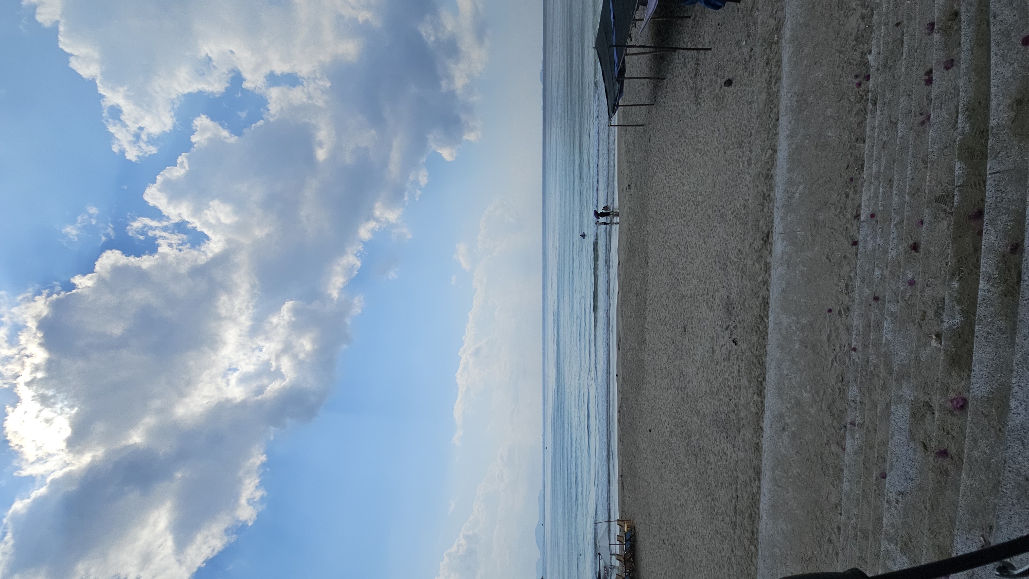 The image shows a beach in Cam Lam, Vietnam. The scene is vertically oriented. On the left side, the sky is visible with scattered clouds and a clear blue sky. In the middle, the ocean extends to the horizon. The right side shows the beach with sand and concrete steps leading down to the sand. There are some beach umbrellas and figures of people walking along the shoreline. The composition is split into thirds, with the sky and clouds on one side, the ocean in the middle, and the beach and stairs on the other.