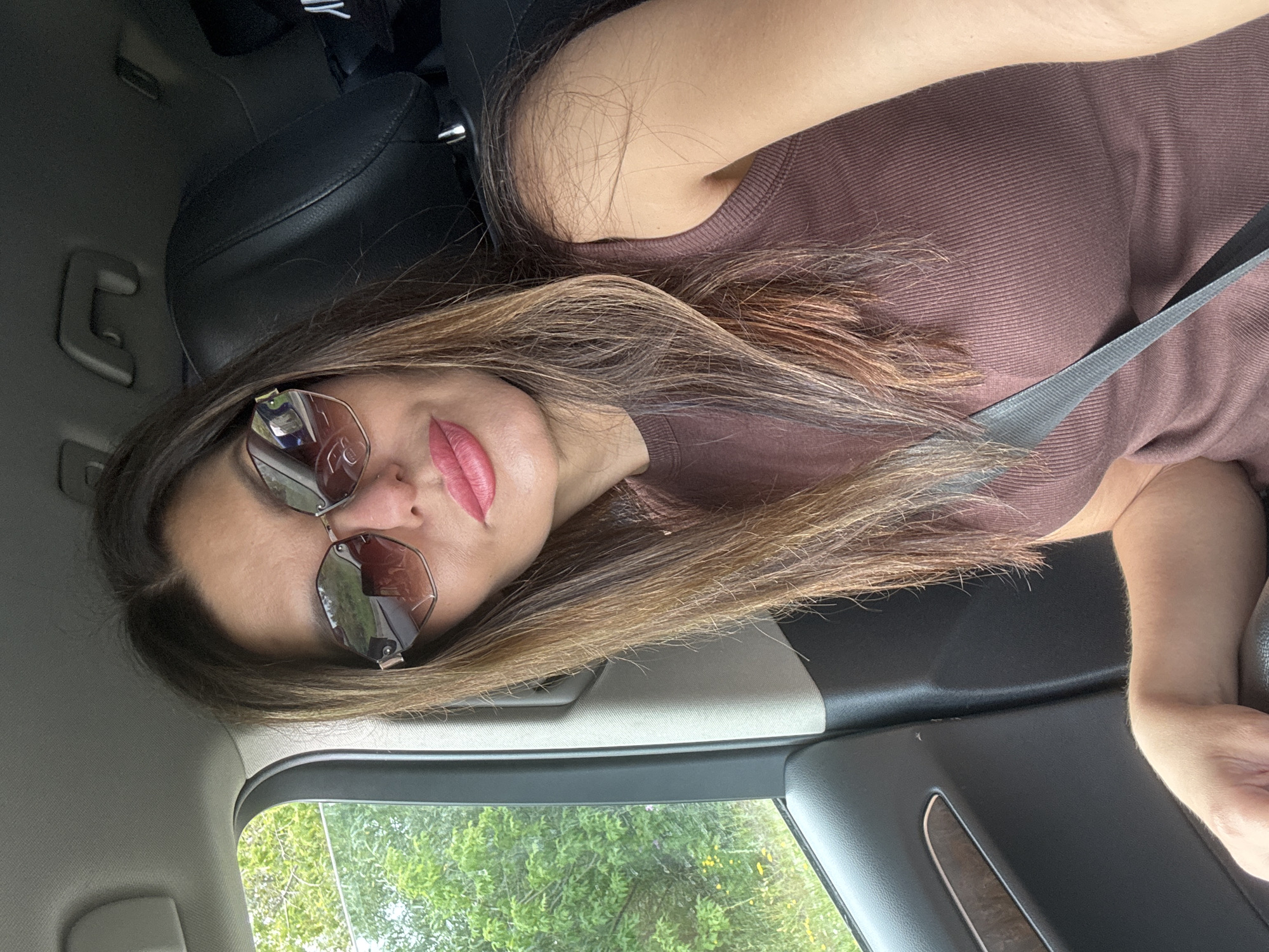 A woman with long brown hair is sitting in the passenger seat of a car. She is wearing sunglasses and a brown sleeveless top. The car interior is visible, including the headrest and a seatbelt. Outside the window, there are trees and greenery. This image was taken on an overcast day. The location is identified as Chaikovsky, Russia.