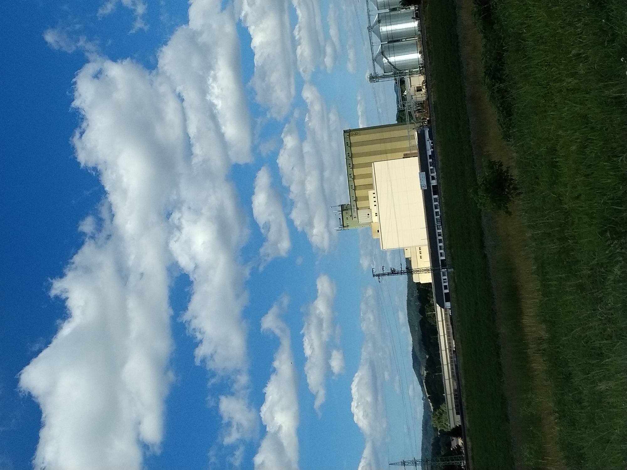 The image depicts an industrial area with several large grain silos and storage facilities, set against a backdrop of rolling green hills under a bright blue sky dotted with white cumulus clouds. A tall, yellow and green building with multiple windows stands prominently, possibly a grain elevator or processing plant. To its right, a row of shiny, silver silos rises, reflecting the sunlight. A lower, cream-colored building with numerous windows extends from the base of the yellow structure. Electrical pylons and power lines are visible throughout the scene, indicating the presence of industrial infrastructure. The foreground features a grassy field with a dark green slope leading up to the industrial buildings. The location is identified as Kapsova Lhota, Czechia. There are no people visible in the image.
