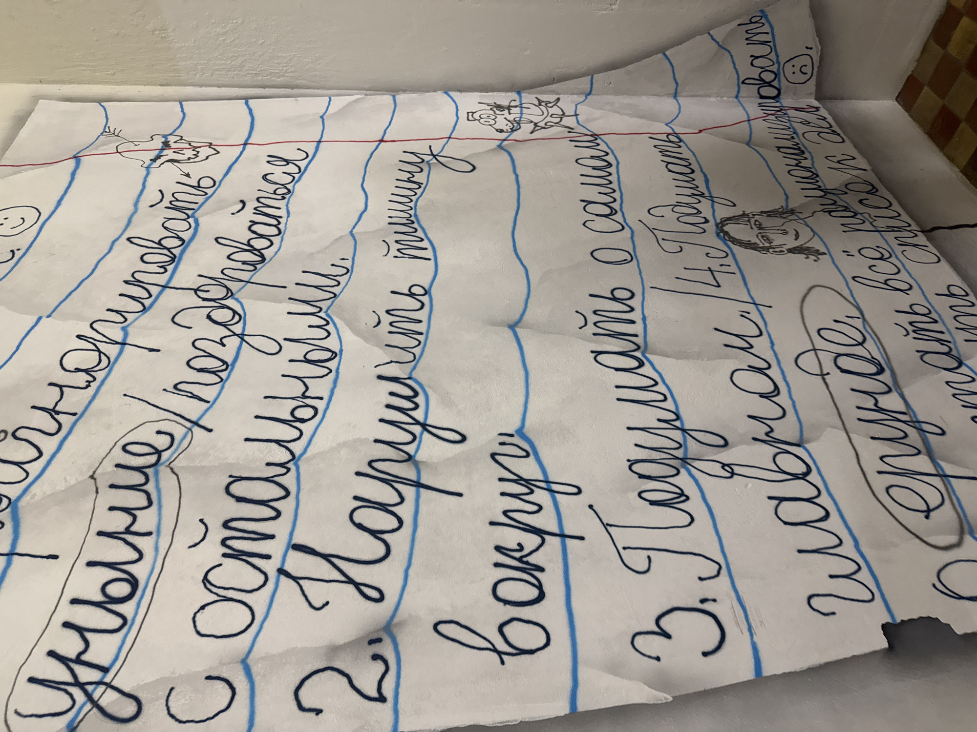 A crumpled piece of lined paper with handwritten Russian text in blue ink is placed on an off-white surface, possibly a table or ledge. The paper is torn along the right side. The blue lines on the paper are typical of notebook paper. The handwritten text appears to be numbered, with each line representing a separate point or instruction. The background behind the paper is blurry, showing a combination of light and dark tones, suggesting a wall or corner. Additionally, there are small cartoonish doodles scattered among the lines of text. The exact location and details of the Moscow setting (if any) cannot be determined from this single image.