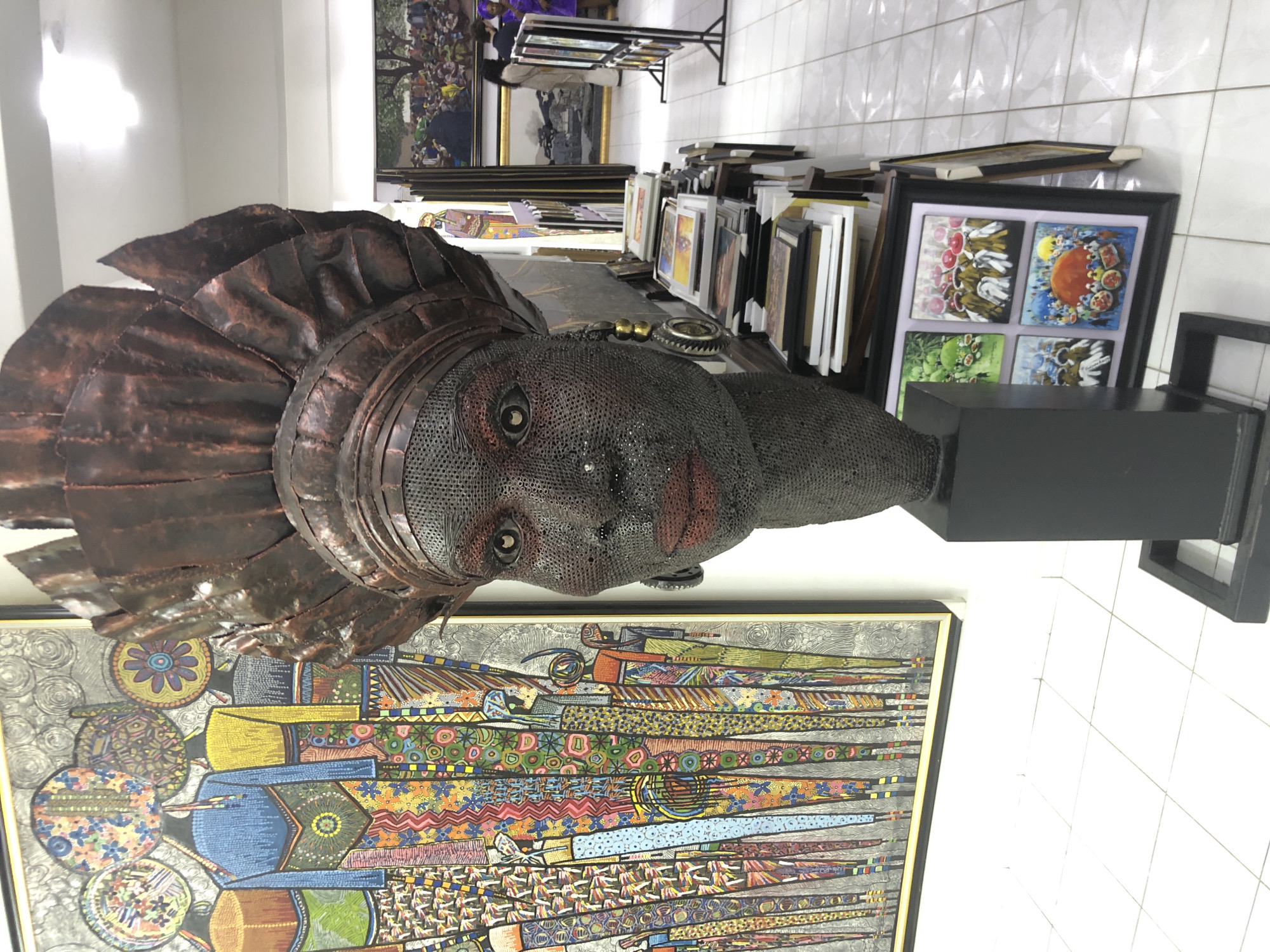 The image depicts a street scene in Alaguntan, Nigeria, focusing on a large, intricate sculpture of a woman's head with a pronounced headdress. The sculpture, made from what appears to be dark metal, has a mesh-like texture for the face and detailed metallic elements for the lips, eyes, and headdress. To the left of the sculpture is a large, colorful painting featuring rows of stylized figures with elaborate patterns on their clothing and circular headdresses. In the background, shelves are stacked with framed artwork, and beyond that, more paintings are visible, some with landscapes and crowd scenes. The floor is tiled with white squares. A few people are discernible in the distance, interacting within the art space, some looking at artworks. The scene suggests an art gallery or a market stall within Alaguntan, showcasing local art and crafts.