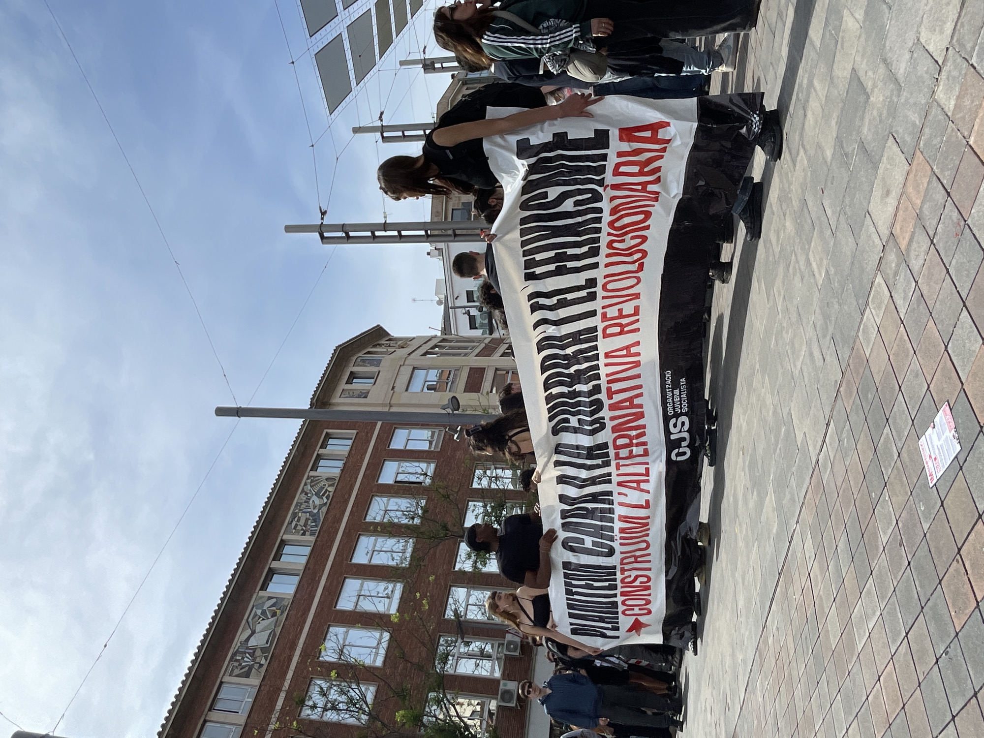A street-level, medium-shot image shows a group of people holding a banner. The banner is white with black and red text, oriented vertically. The text appears to be in Spanish/Catalan and includes the word "feminism." The banner also features the acronym "CJS," presumably an organization abbreviation. People of various ages and ethnicities are holding the banner. They are dressed in casual clothing.
The background features a brick building with multiple windows and decorative panels near the top. The sky is light blue with some clouds. Overhead wires and poles are visible, suggesting an urban setting. The ground is paved with square stones. Based on the architectural style and language on the banner, the location is likely Barcelona, Spain.