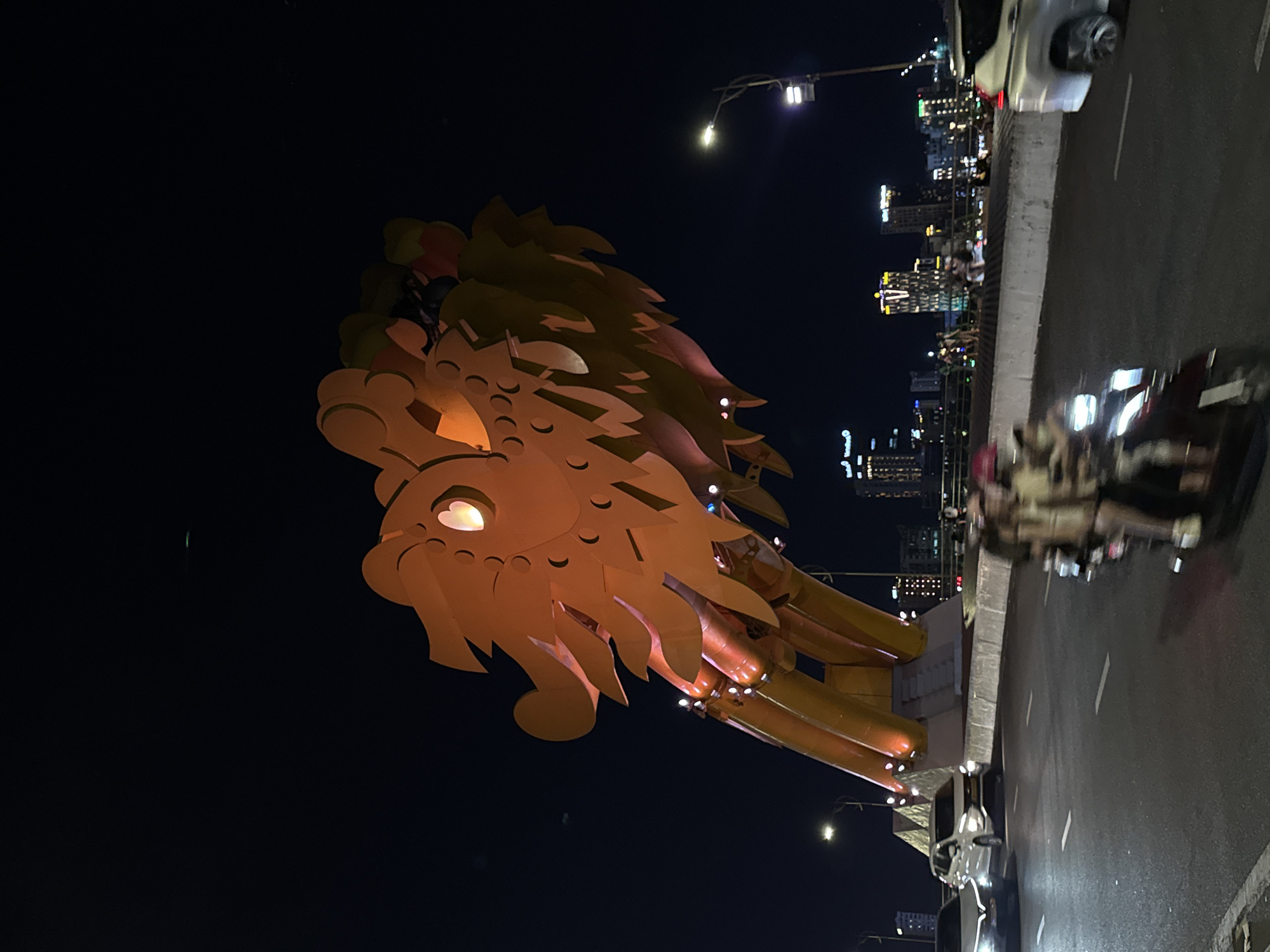 The image captures the Dragon Bridge in Da Nang, Vietnam at night. The bridge's iconic dragon head, illuminated in a warm orange hue, dominates the upper portion of the frame. Below, a street scene unfolds with vehicles on the road. Two cars are visible in the foreground, one white and one silver. A motorcycle with two passengers, blurred by motion, travels in the right lane. In the background, the cityscape of Da Nang rises with illuminated buildings and streetlights. A few individuals can be seen on a walkway or bridge railing in the distance, looking out at the view. The dark night sky provides a stark contrast to the illuminated structures and vehicles, highlighting the vibrant atmosphere of the city at night.