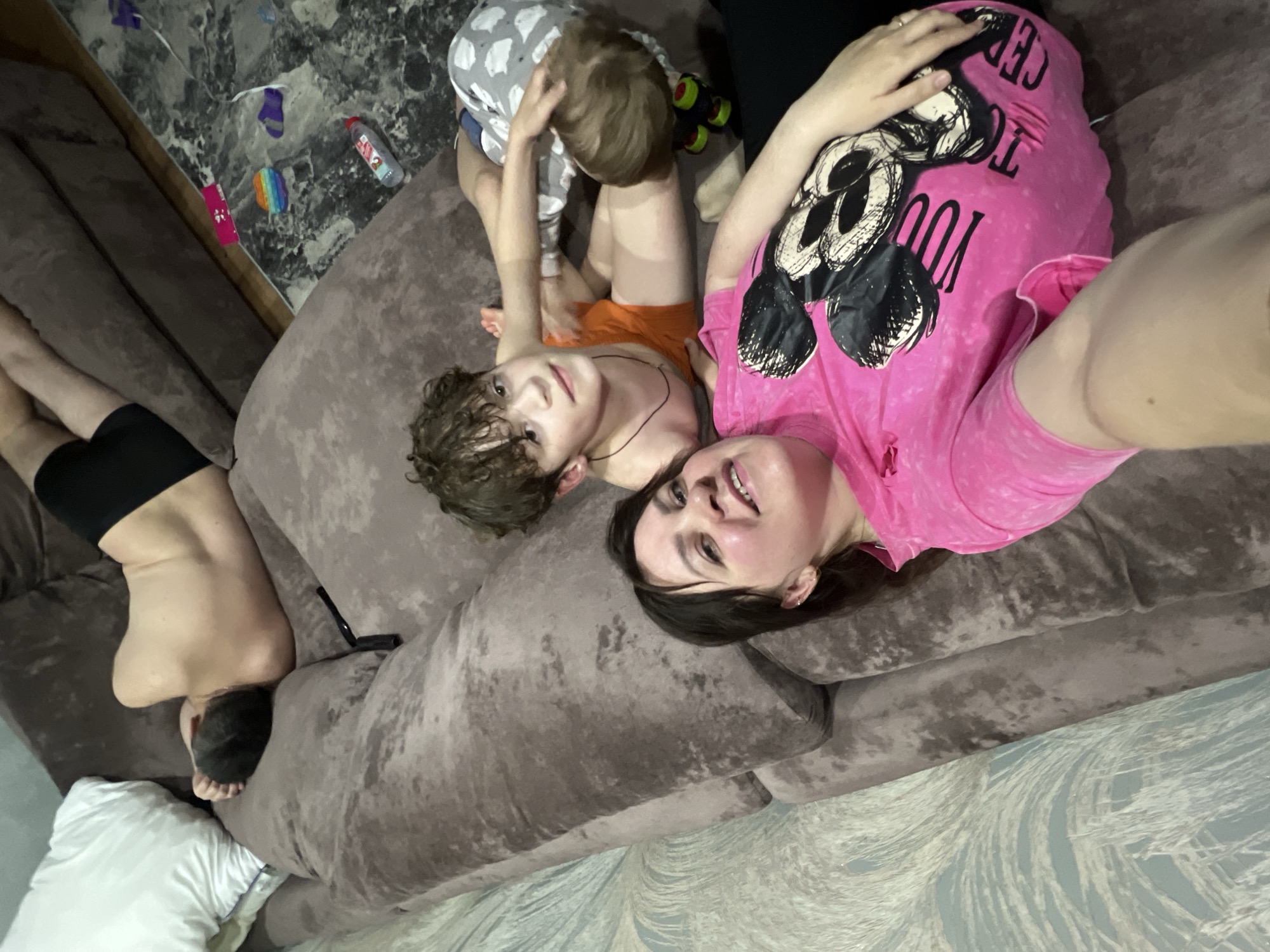 A selfie shows a woman in a pink Mickey Mouse shirt lying on a gray couch with three children. One child is prone with his legs on the couch. Another child is looking at the camera while wearing an orange shirt. A third, younger child with a grey patterned onesie stands near them. A table with toys and a bottle sits next to the couch. The couch is in front of a light blue patterned floor, presumably inside a home. The location in the city of Noviy, Russia, cannot be determined from the image.
