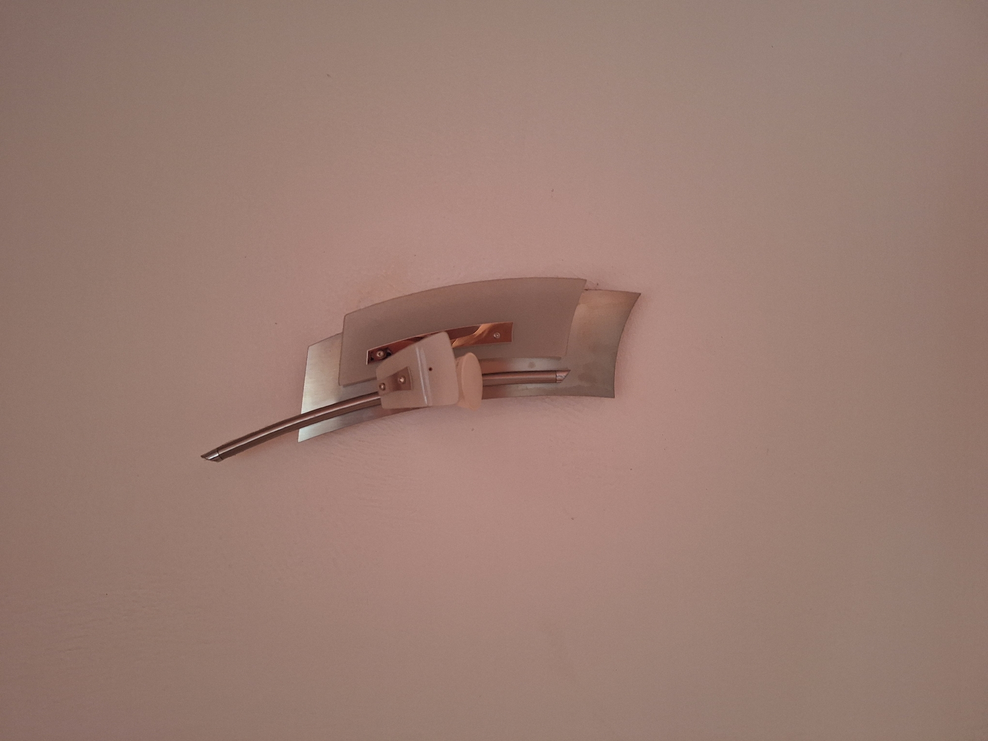 An outdoor wall-mounted light fixture is affixed to a textured, light pink wall. The fixture has a metallic, abstract design with overlapping panels and a slender, pointed element extending upwards. There are no people or animals visible in the image. The overall scene appears to be a residential or commercial exterior. The location in Varna, Bulgaria, is not discernible from the visual information provided.