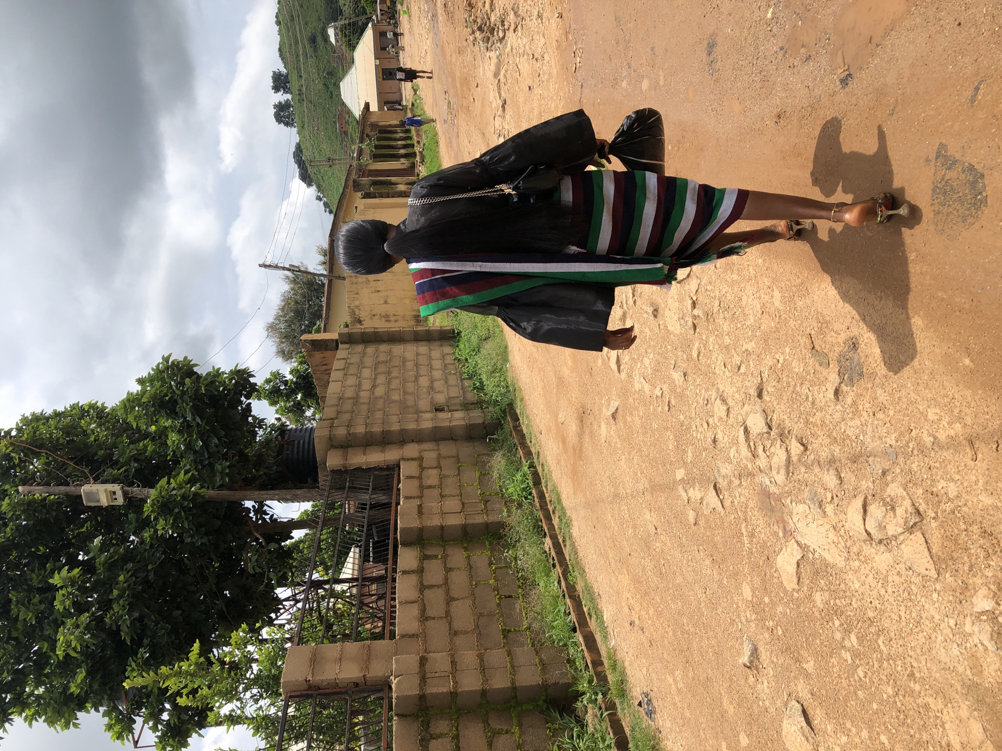 The image depicts a woman walking away from the camera on a dusty, dirt road in Jos, Nigeria. She has long, dark hair pulled back into a ponytail and is wearing a black top with wide, flowing sleeves and a horizontally striped skirt in shades of green, white, and maroon. A colorful sash is draped over her shoulders, with stripes matching those of her skirt. She carries a black plastic bag in her right hand and wears high-heeled sandals.
The road is uneven and appears to be recently rained on in places, with puddles visible. To the left of the road is a low wall made of tan concrete blocks, overgrown with grass and moss. Behind the wall, there is greenery and what appears to be a metal fence. Further down the road, more buildings are visible, with a hillside in the background under a cloudy sky. The overall environment suggests a rural or semi-urban setting.
