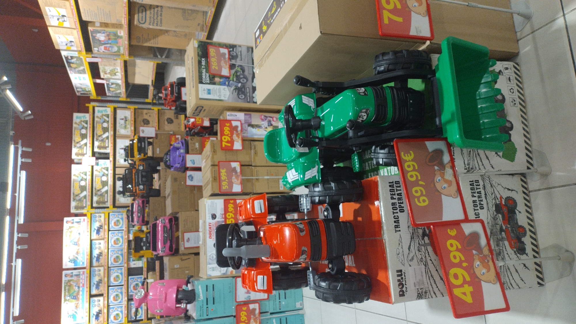 In a retail store in Pylaia Municipal Unit, Greece, a display of children's ride-on toys is presented. The foreground features two pedal-powered tractors, one orange and one green, positioned on top of their respective product boxes. The orange tractor is on the left, and the green tractor is on the right. Red signage with yellow lettering indicating prices of "49,99 €" and "69,99 €" is visible in front of the tractors. Behind these, a variety of other ride-on vehicles, including pink scooters and cars, as well as larger toy trucks and ATVs, are displayed on yellow shelving units that extend towards the back of the store. The environment appears to be a well-lit toy section within a larger retail establishment, with numerous cardboard boxes stacked on shelves.