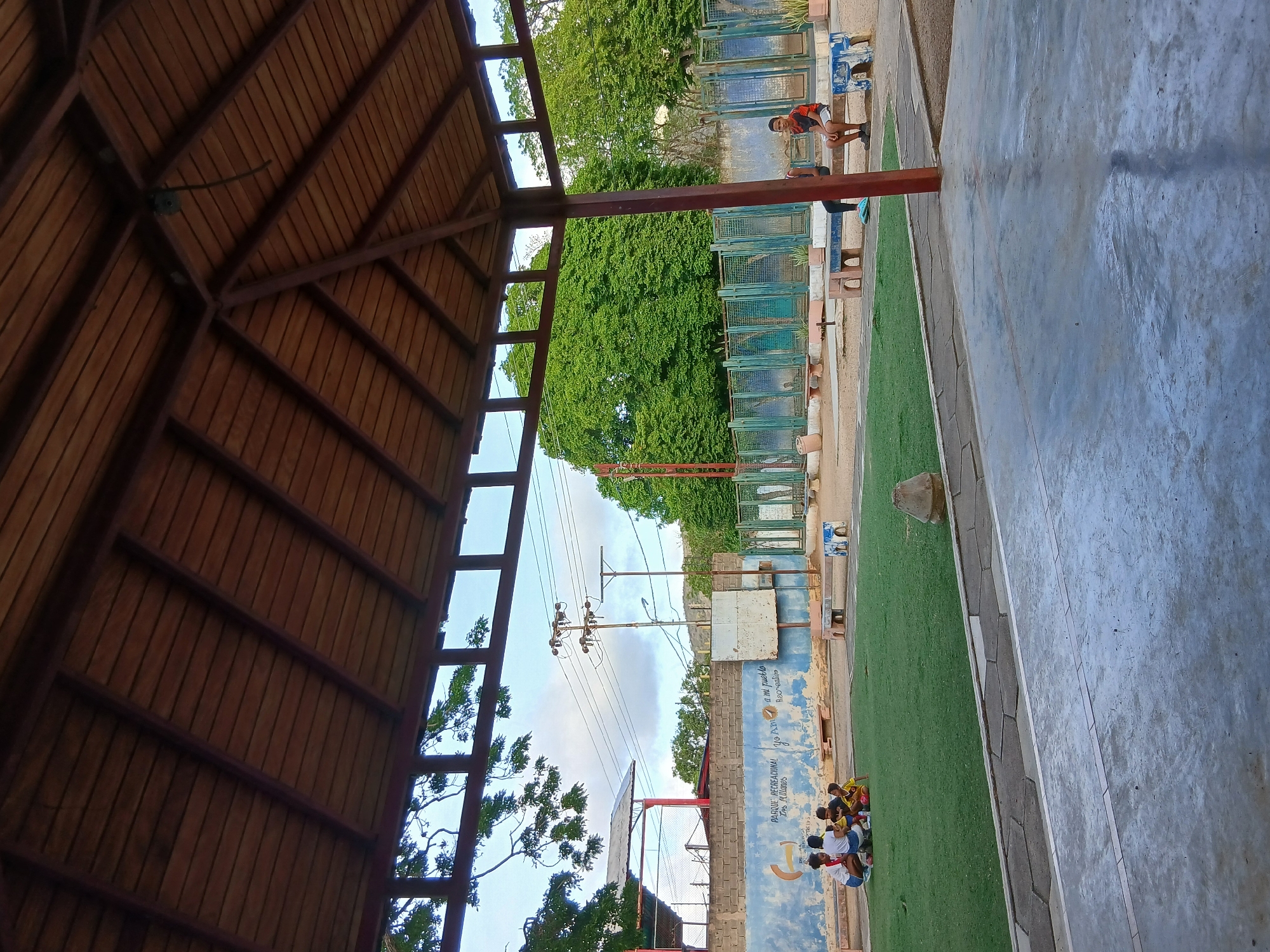 A group of young children are gathered on a patch of artificial turf under a wooden-roofed pavilion in a park. The children are mostly facing each other, engaged in an activity or conversation. In the background, there is a blue and white mural on a brick wall, with text that appears to be in Spanish, potentially indicating the park's name, "PARQUE RECREACIONAL LOS MILANES." There are several metal fences and benches visible in the middle ground, and a large tree with dense green foliage dominates the background. A lone young male sits on a bench to the right, facing away from the children. The scene appears to be an outdoor recreational area, likely in a warm climate given the vegetation. The overall atmosphere is casual and communal. The image is taken from within the pavilion, looking out towards the park. The graffiti on the wall suggests the location is in Juan Griego, Venezuela.