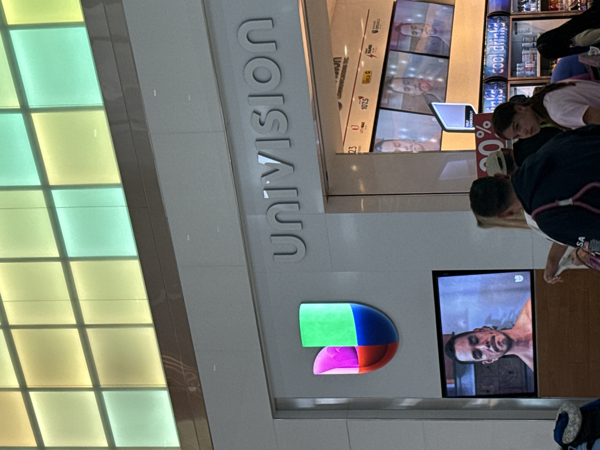 The image shows the interior of a store with the "UNIVISION" logo prominently displayed. Adjacent to the logo is a television screen playing content. People are present in the store, interacting with the displays and potentially shopping. Drinks are visible in the background. The image shows the terminal at the Fort Lauderdale-Hollywood International Airport (FLL).