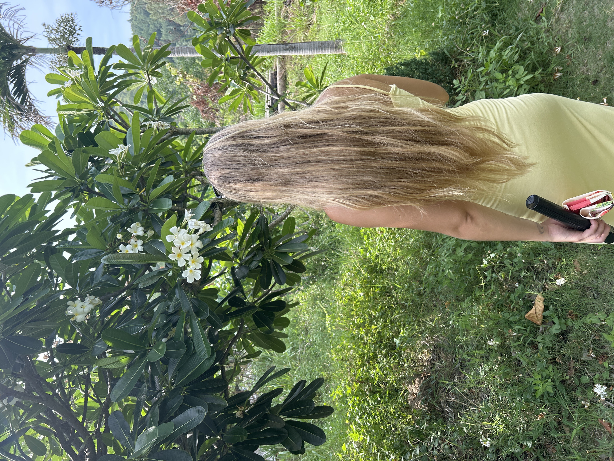 A woman with long blonde hair, wearing a light yellow dress, stands with her back to the camera, facing a lush green outdoor area. She is holding a black object and a folded pink and white item in her right hand. The environment appears to be a tropical garden, with a plumeria tree in the foreground adorned with white flowers and dense green foliage. Behind the tree, a palm tree and more greenery are visible, suggesting a location in a warm climate. The ground is covered in grass and small plants, with some fallen leaves scattered around. The scene is lit by natural daylight, casting soft shadows. The location is identified as Nam Nha Trang Ward, Vietnam.