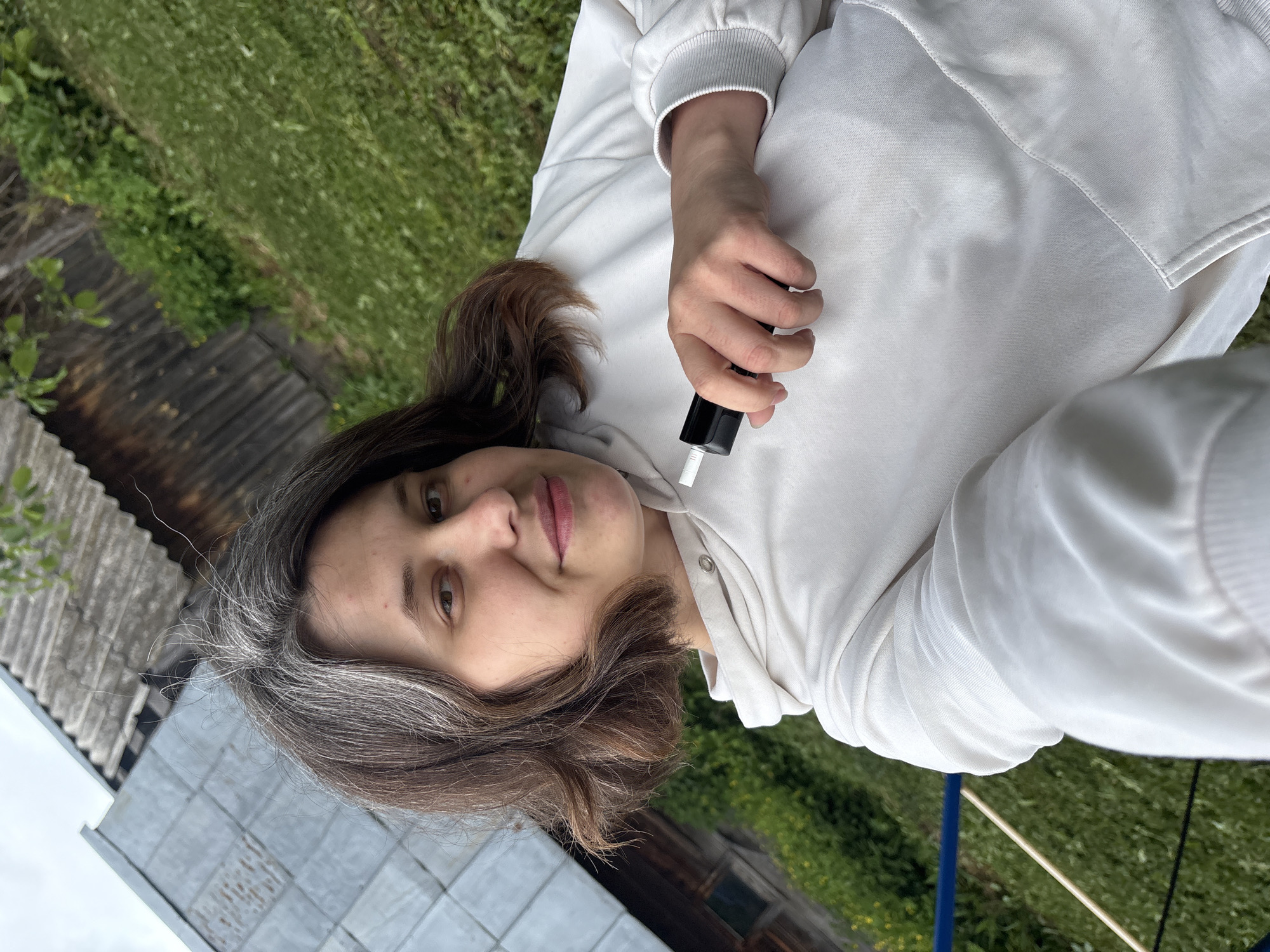 An adult female, appearing in her late 20s to early 40s, is depicted in a close-up selfie taken outdoors in the City of Фоки, Russia. She has dark brown, shoulder-length hair with visible grey streaks, styled with a slight wave. Her complexion is fair, and she has brown eyes, a subtle smile, and light blemishes on her forehead. She is wearing a white hooded sweatshirt with ribbed cuffs. Her right hand holds a black electronic device, possibly a vape pen or electronic cigarette, with a white mouthpiece. The background consists of a sloped grassy area with patches of yellow flowers and green foliage. To the left, a building with a grey corrugated metal roof is visible. To the right, part of a wooden structure, possibly a house or barn, with a dark corrugated roof, is present. A blue horizontal bar and a wooden pole are partially visible in the lower-left foreground. The lighting suggests daylight conditions.
